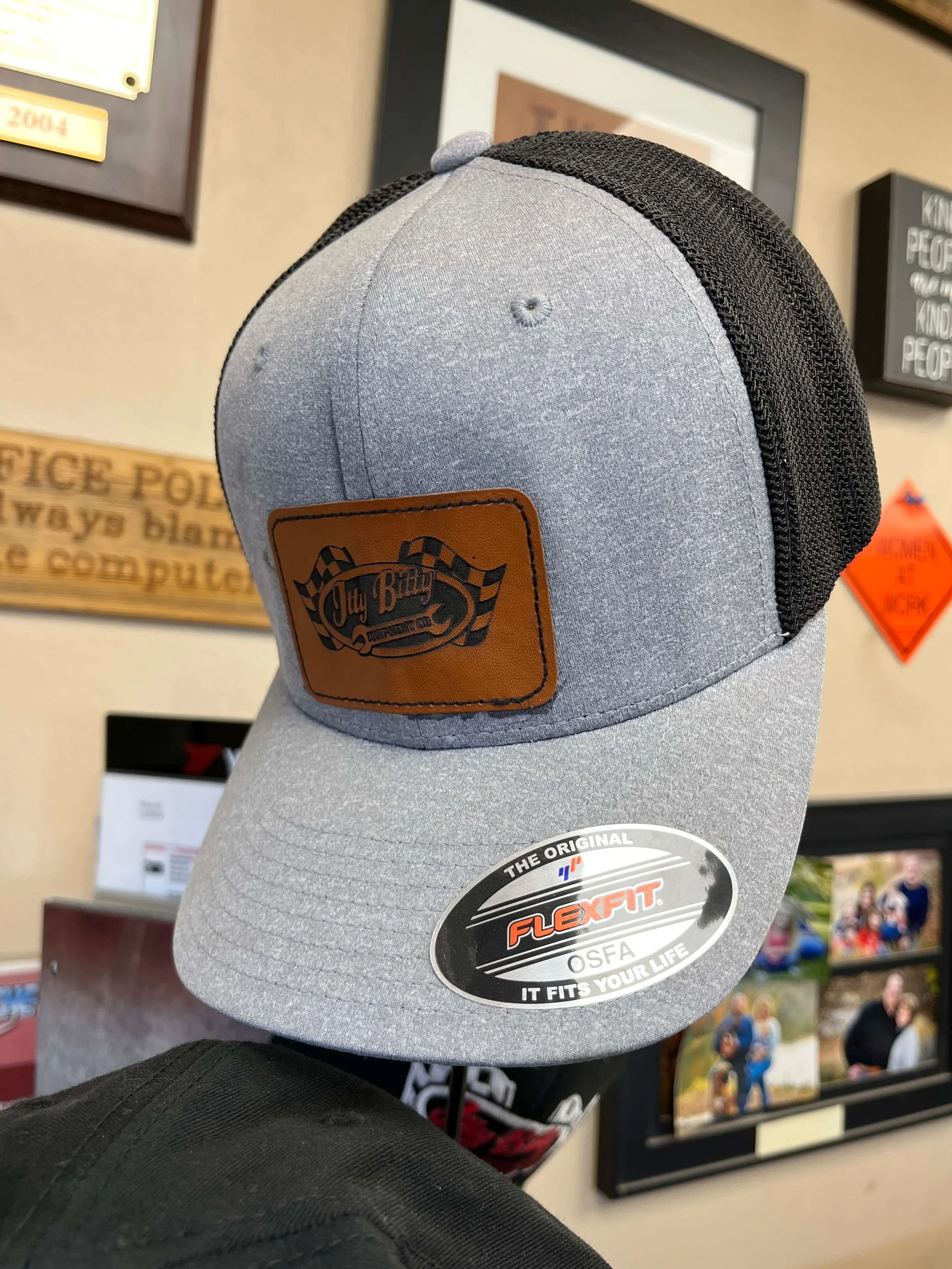 Gray and black trucker hat with "Itty Bitty" logo and "Flexfit" sticker displayed, against a background with framed photos and signs.