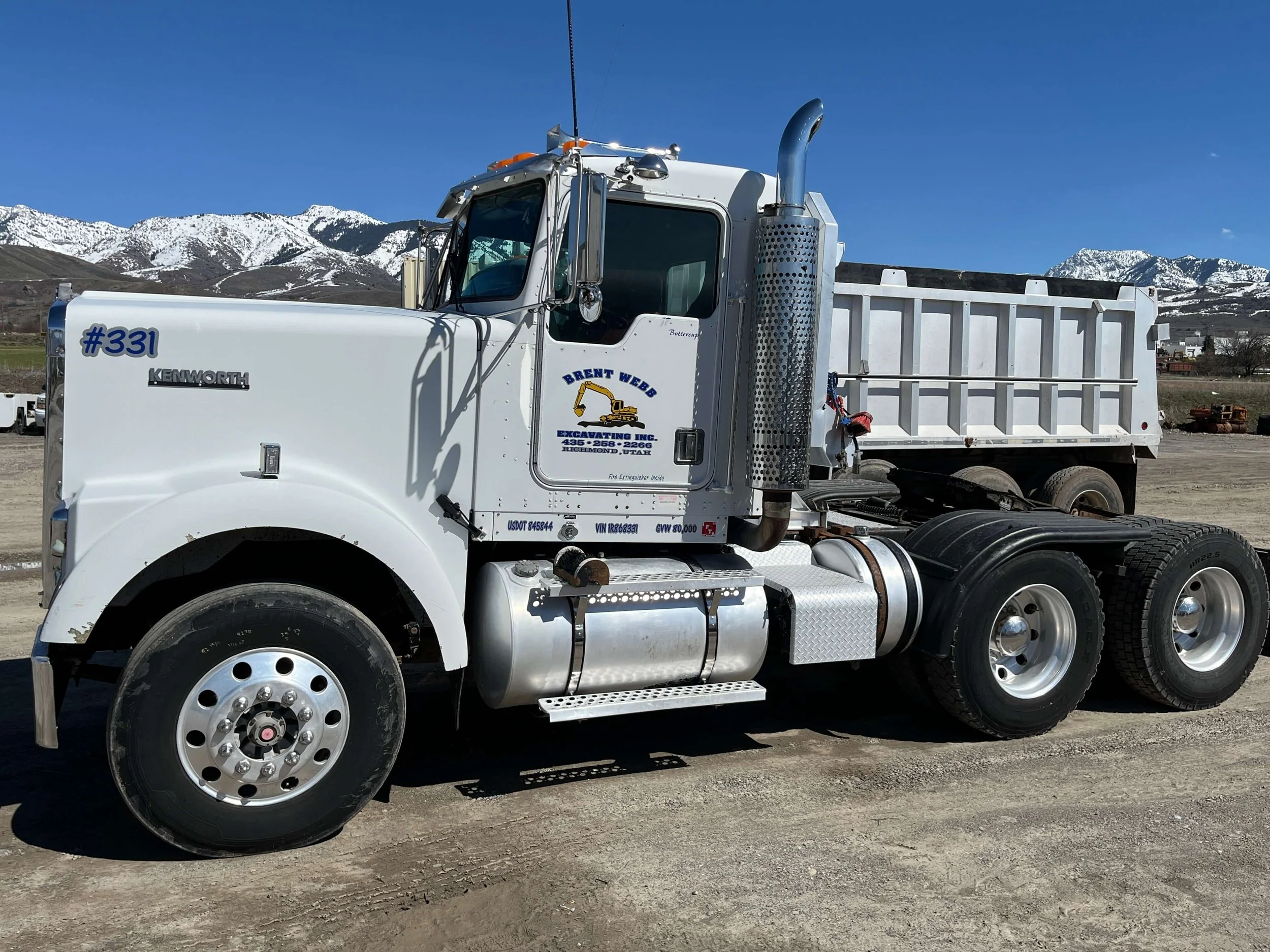 White Kenworth dump truck parked on dirt ground with snow-covered mountains in the background.
