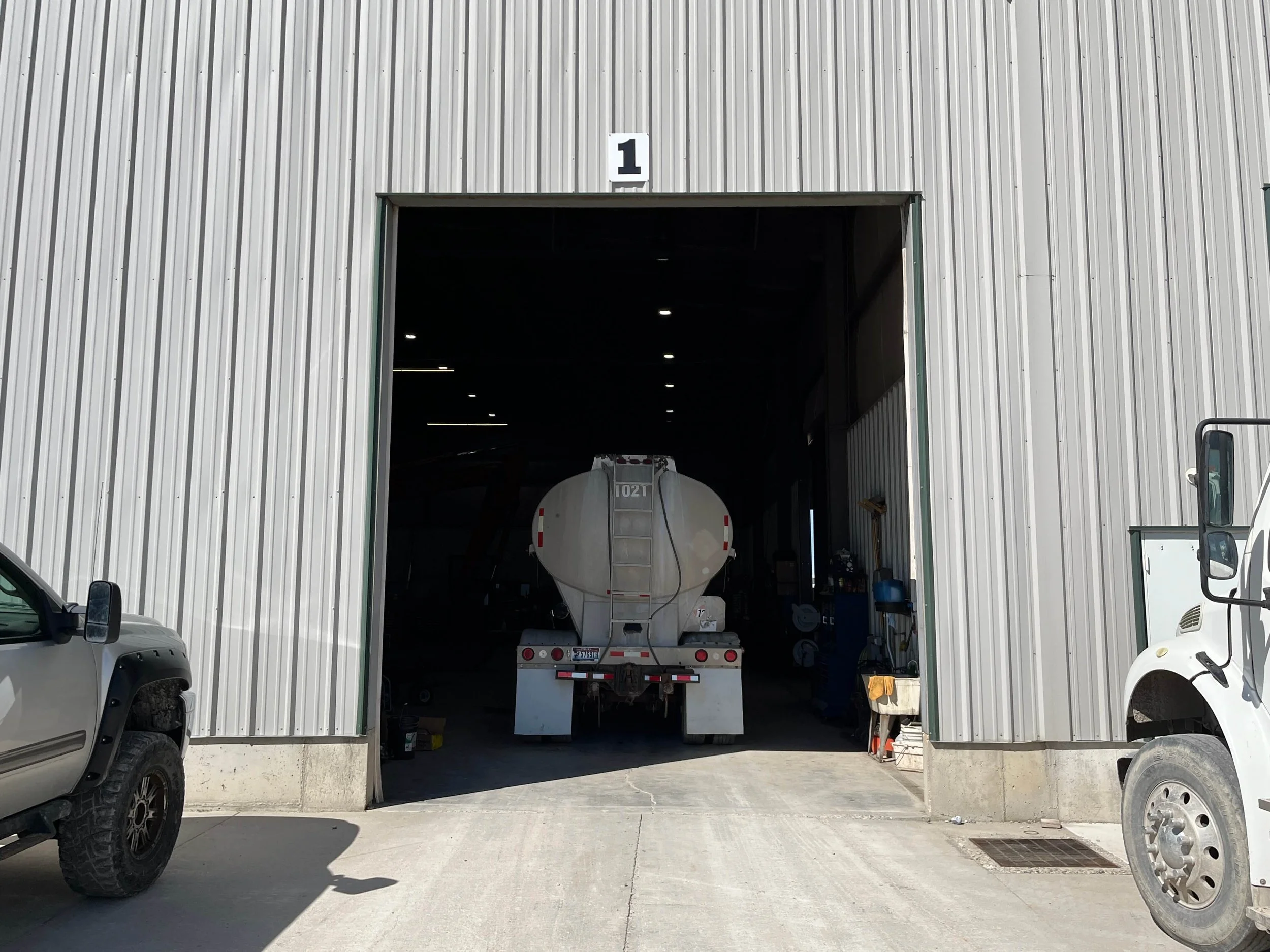 Entrance of a warehouse or garage with a tanker truck partially inside. Two other trucks are parked near the entrance, which is labeled with a number one above. The area is surrounded by a corrugated metal exterior.
