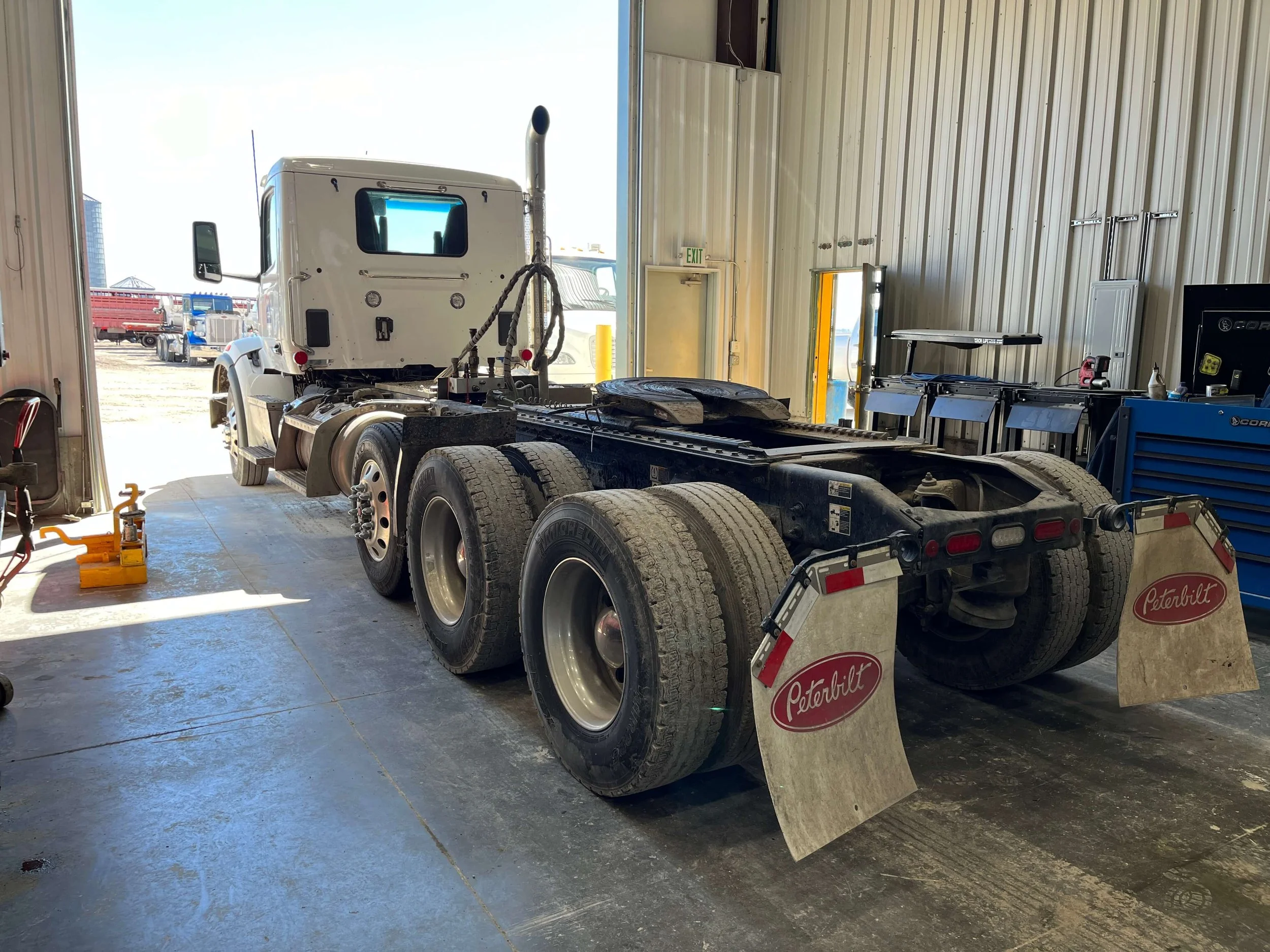 Truck inside a workshop with a Peterbilt logo on mud flaps, showing the rear view with dual wheels and a partially visible cab.