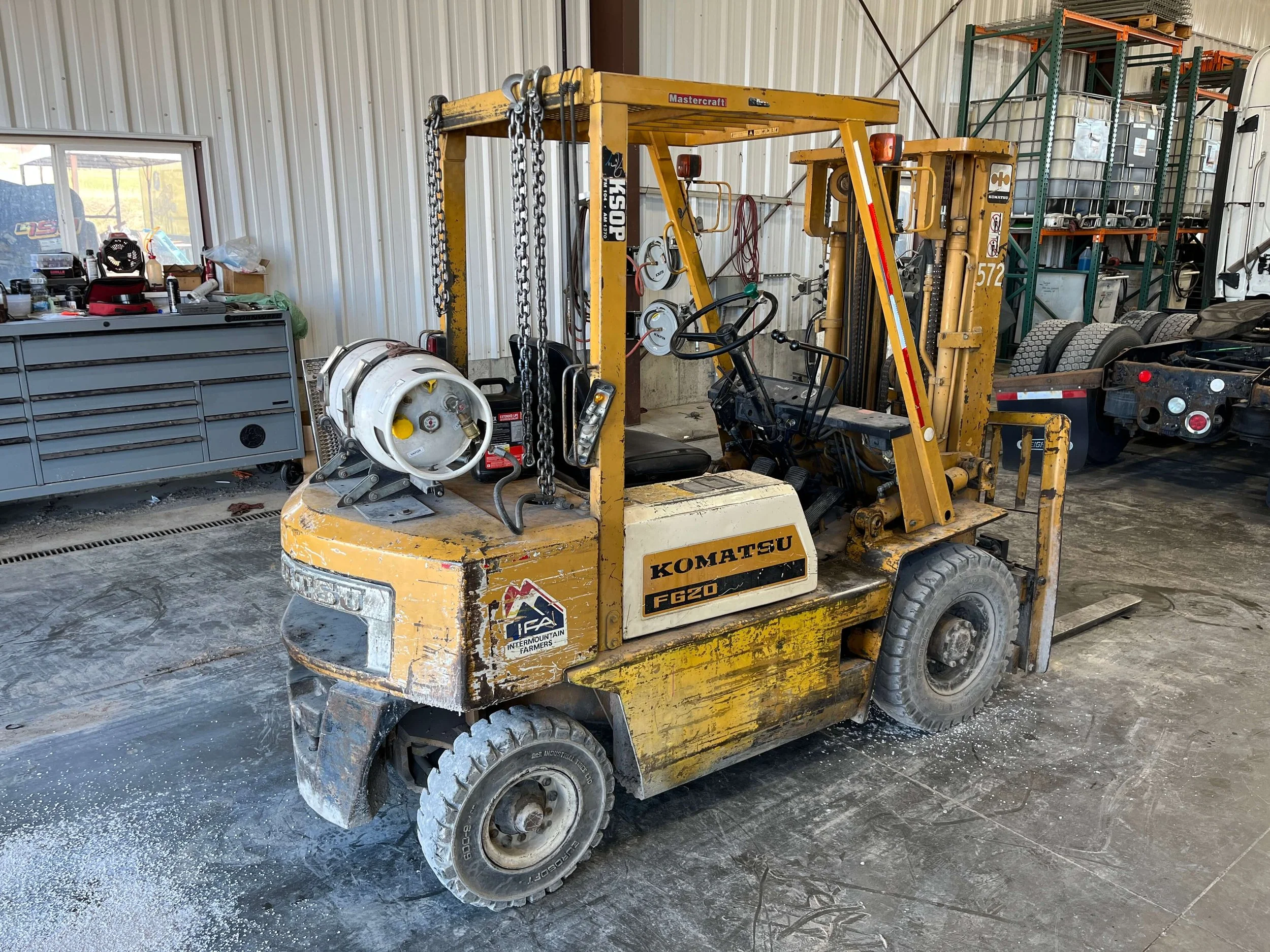 Yellow Komatsu FG20 forklift inside a workshop, with a propane tank attached to the rear. The workshop contains shelves, a toolbox, and a truck chassis in the background.