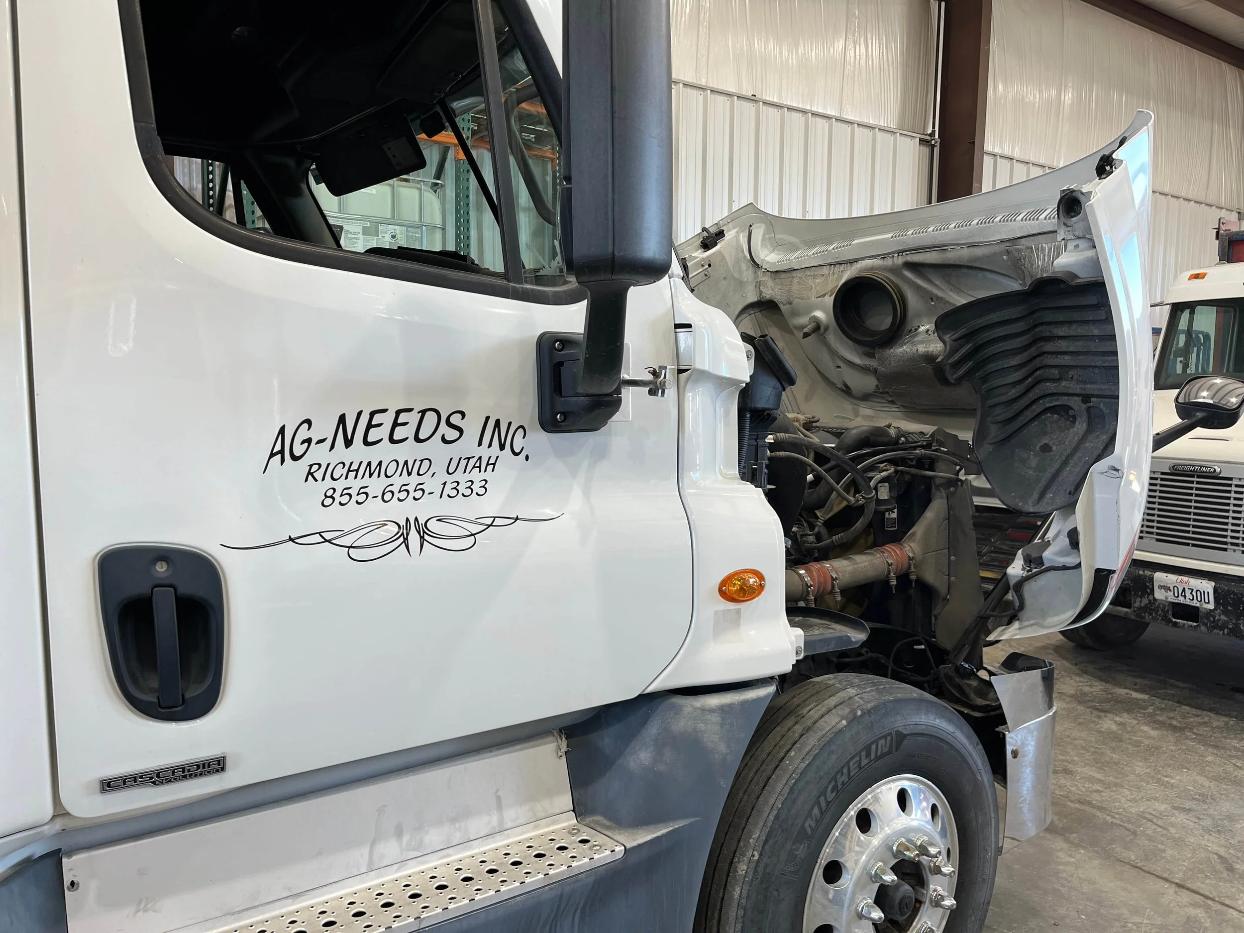 Semi-truck with hood open displaying AG-NEEDS INC. logo, Richmond, Utah location, and contact number, in a garage setting.