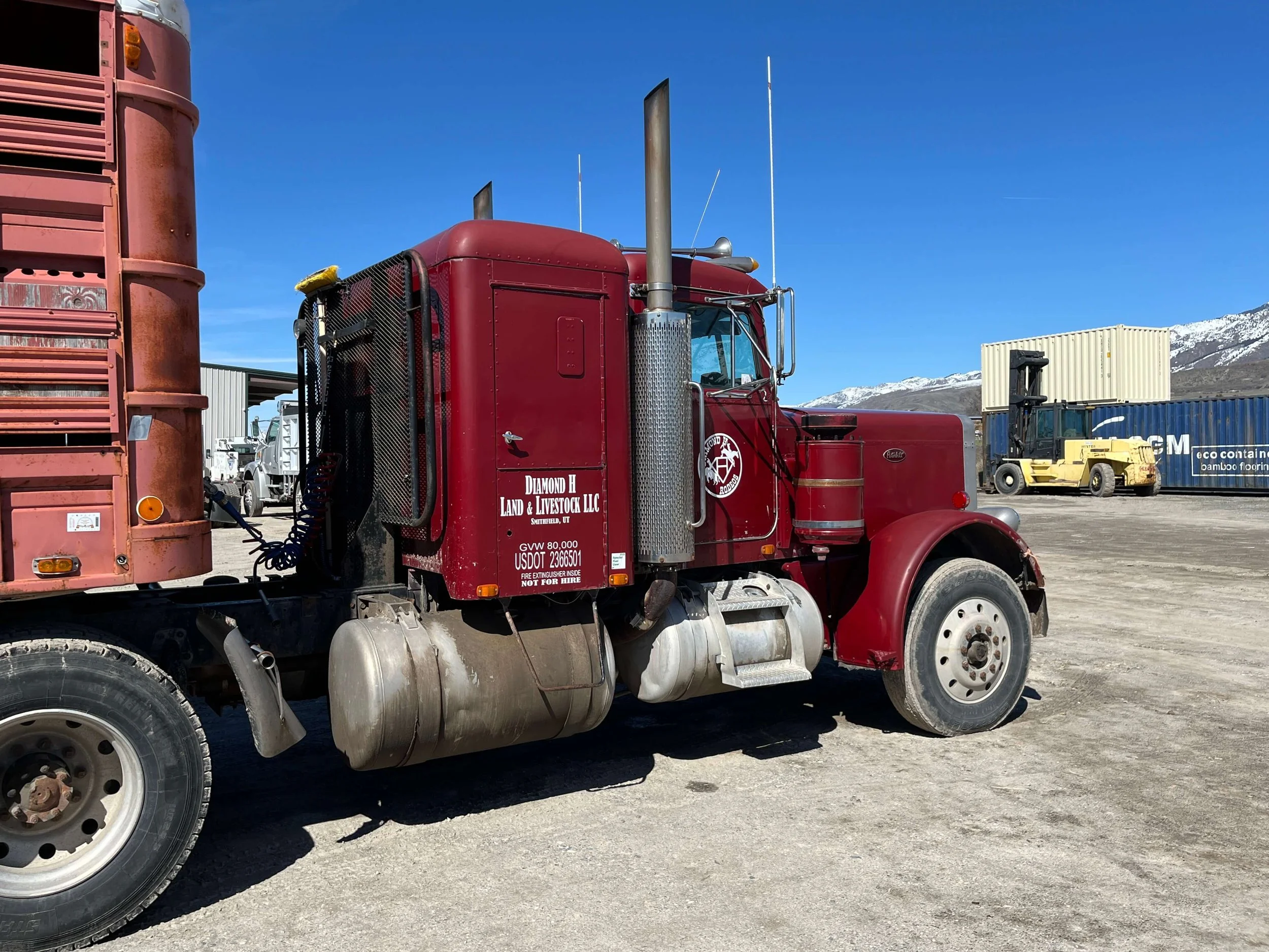 Red semi-truck parked in an industrial area with livestock trailer. The truck has the logo and text "Diamond H Land & Livestock LLC" on the door. Industrial equipment and shipping containers are in the background, with a clear blue sky overhead.