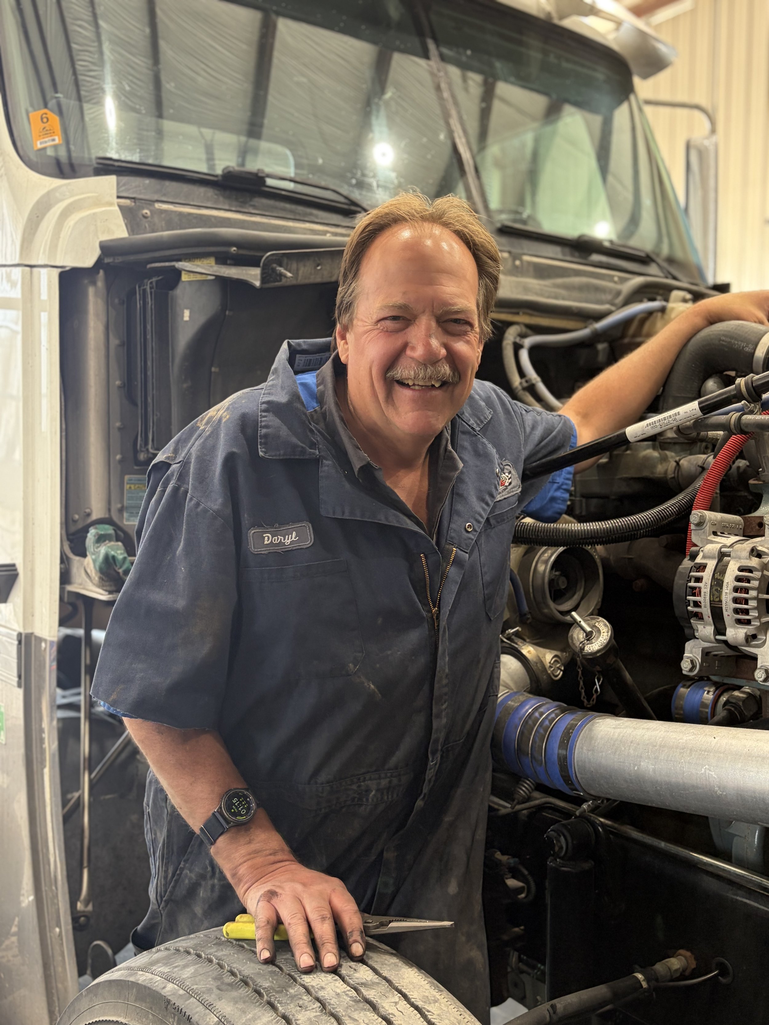 Daryle Black working in a garage, standing near a utility sink inside a workshop. Nearby are NAPA auto parts storage cabinets with tools. In the background, gas trucks are visible outside, and there is a large silo in the distance.