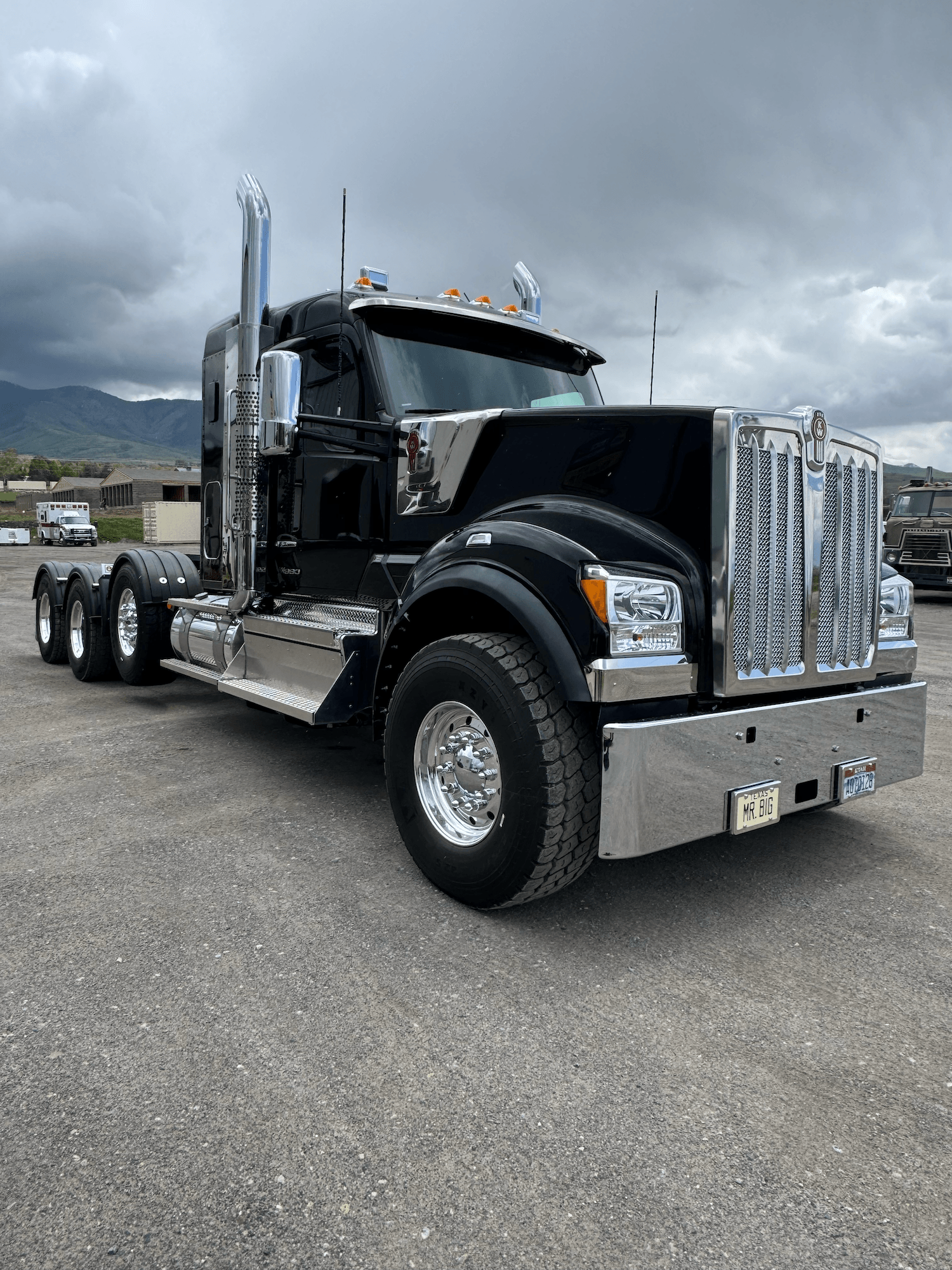 Black semi-truck with chrome details parked on a gravel lot under cloudy sky, mountains in the background.