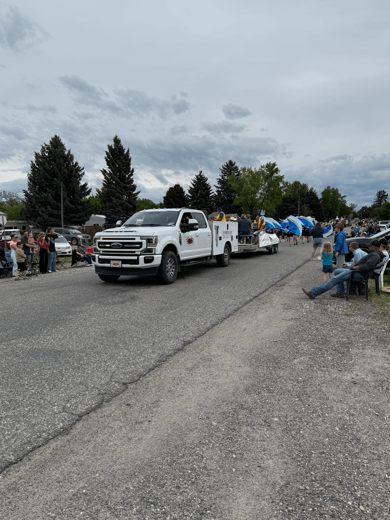 A parade with people sitting on the sidewalk and on chairs, watching a float with band members holding umbrellas, moving down a street lined with trees and onlookers.