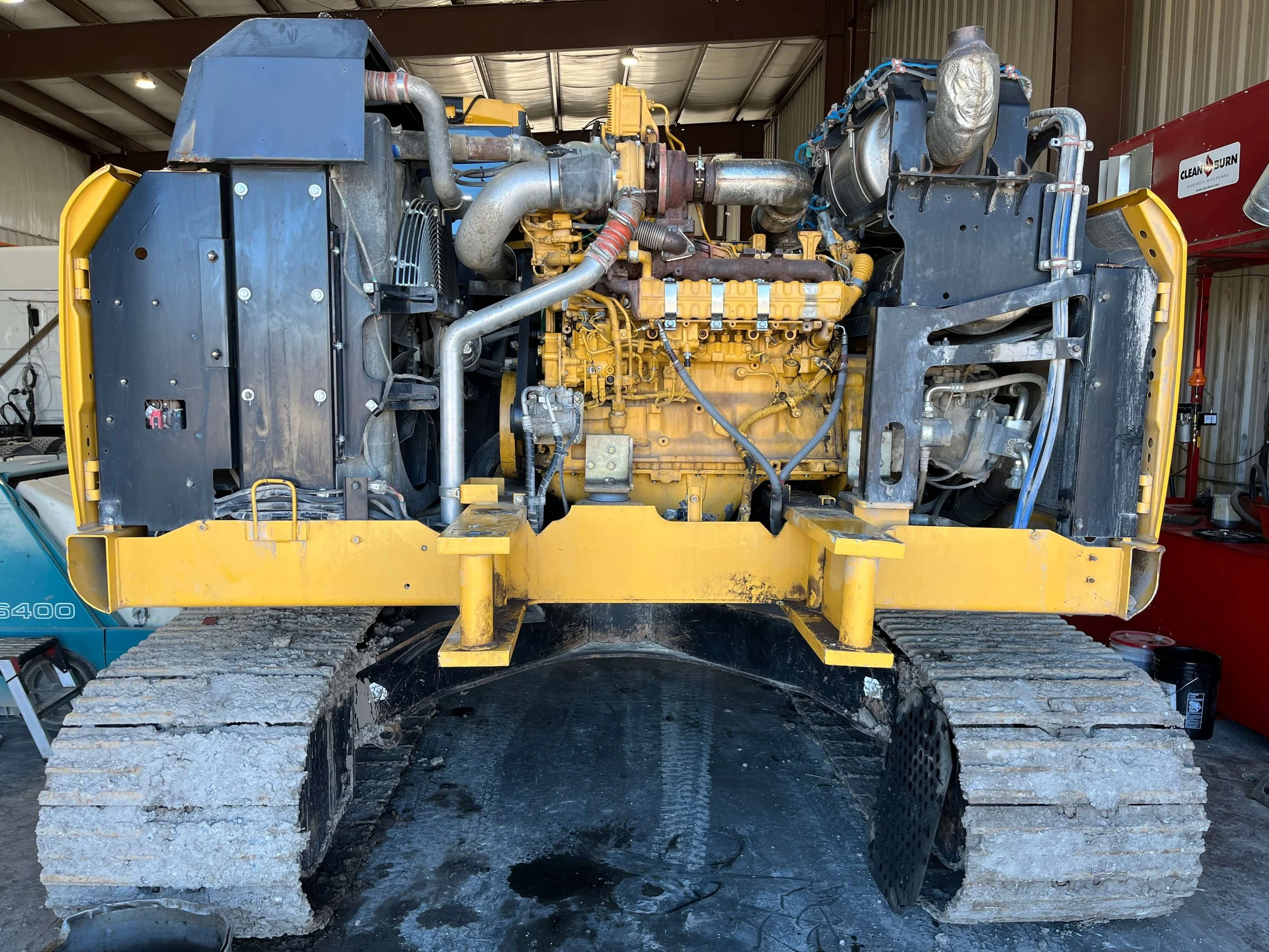 Heavy machinery engine with exposed components, including a yellow engine block, metal pipes, and rubber tracks, inside a warehouse setting.