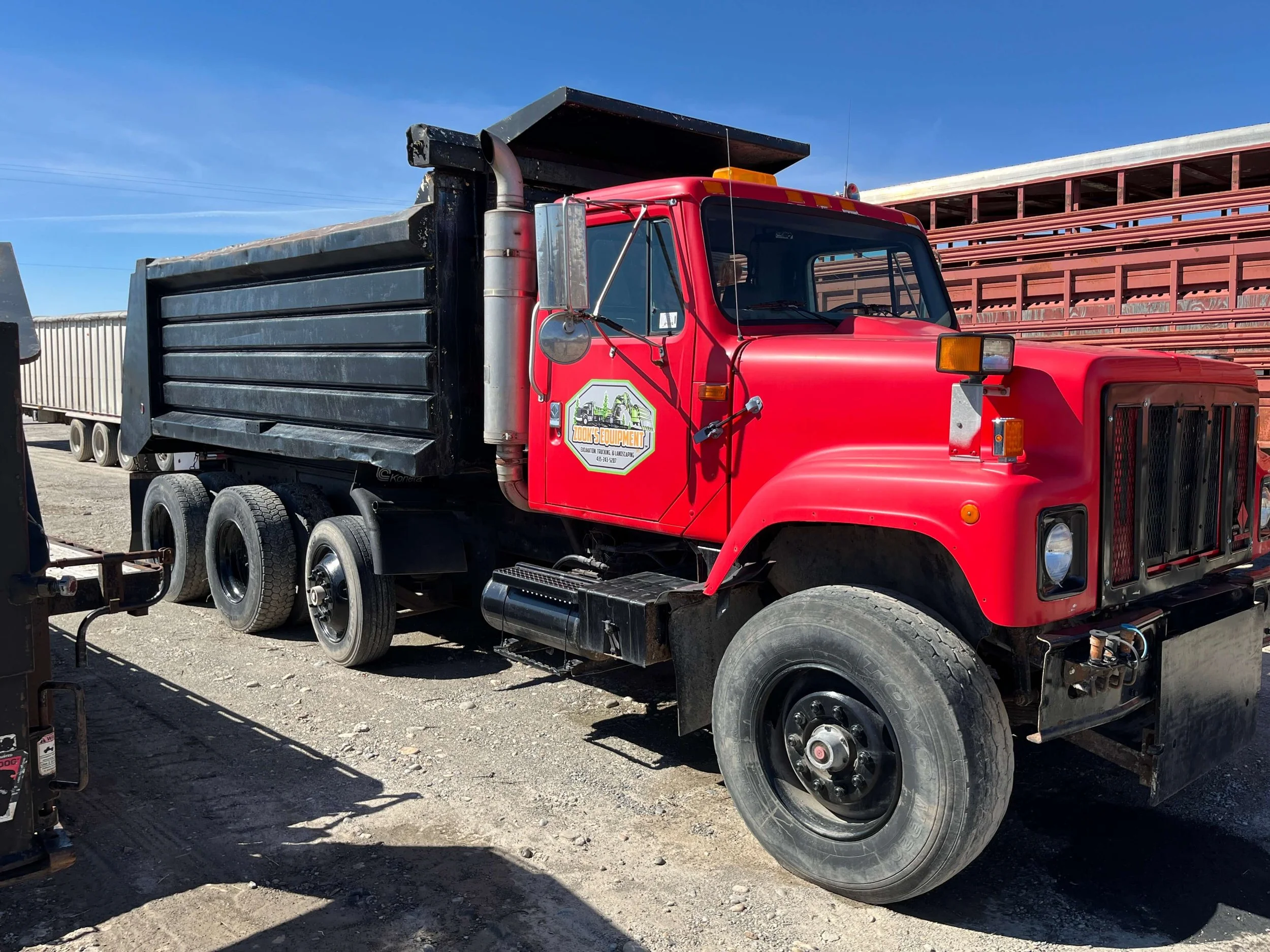 A red dump truck parked on a dirt lot under a clear blue sky, with a black dump bed and multiple axles. A logo is visible on the side of the truck.