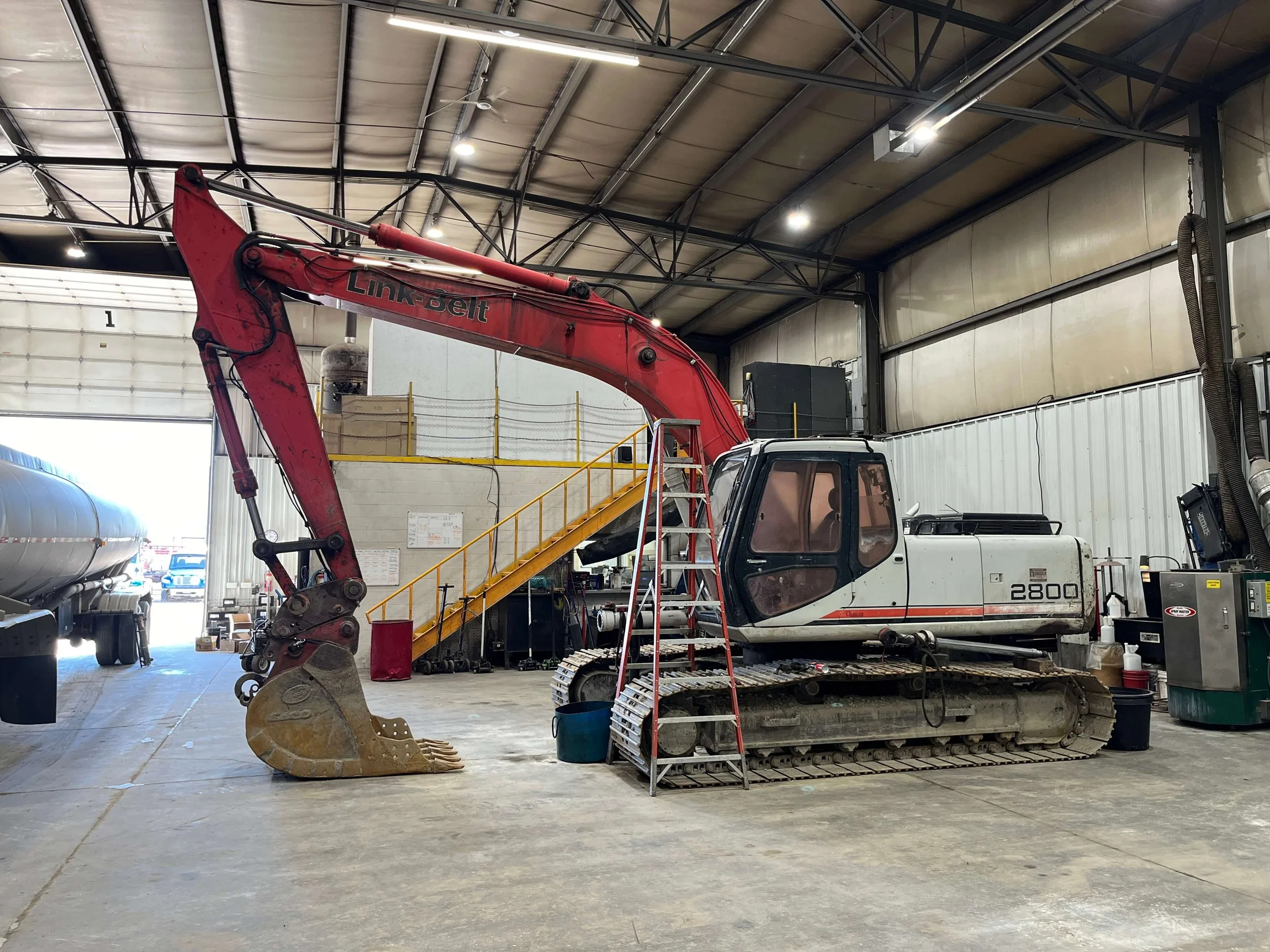 Excavator in a warehouse with an industrial setting, featuring a ladder leaning against it.