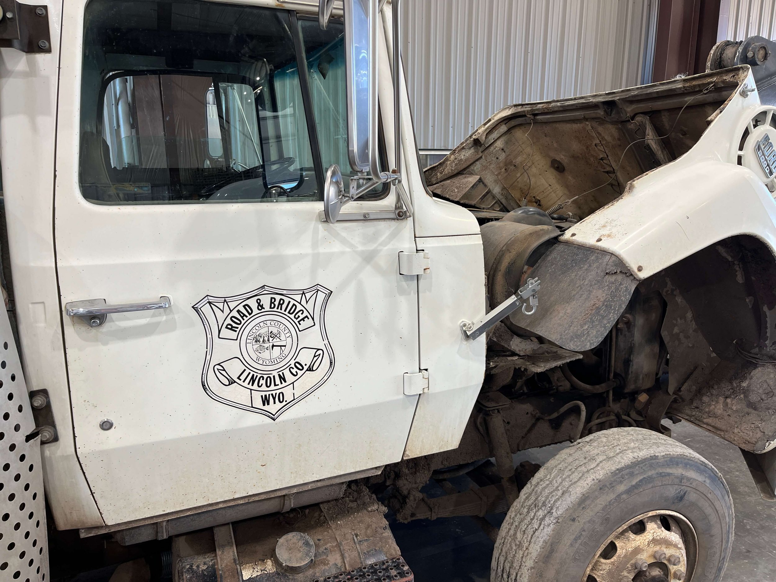 An old white truck in a garage with "Road & Bridge Lincoln Co. Wyo." written on the door and its hood open, revealing mechanical parts.
