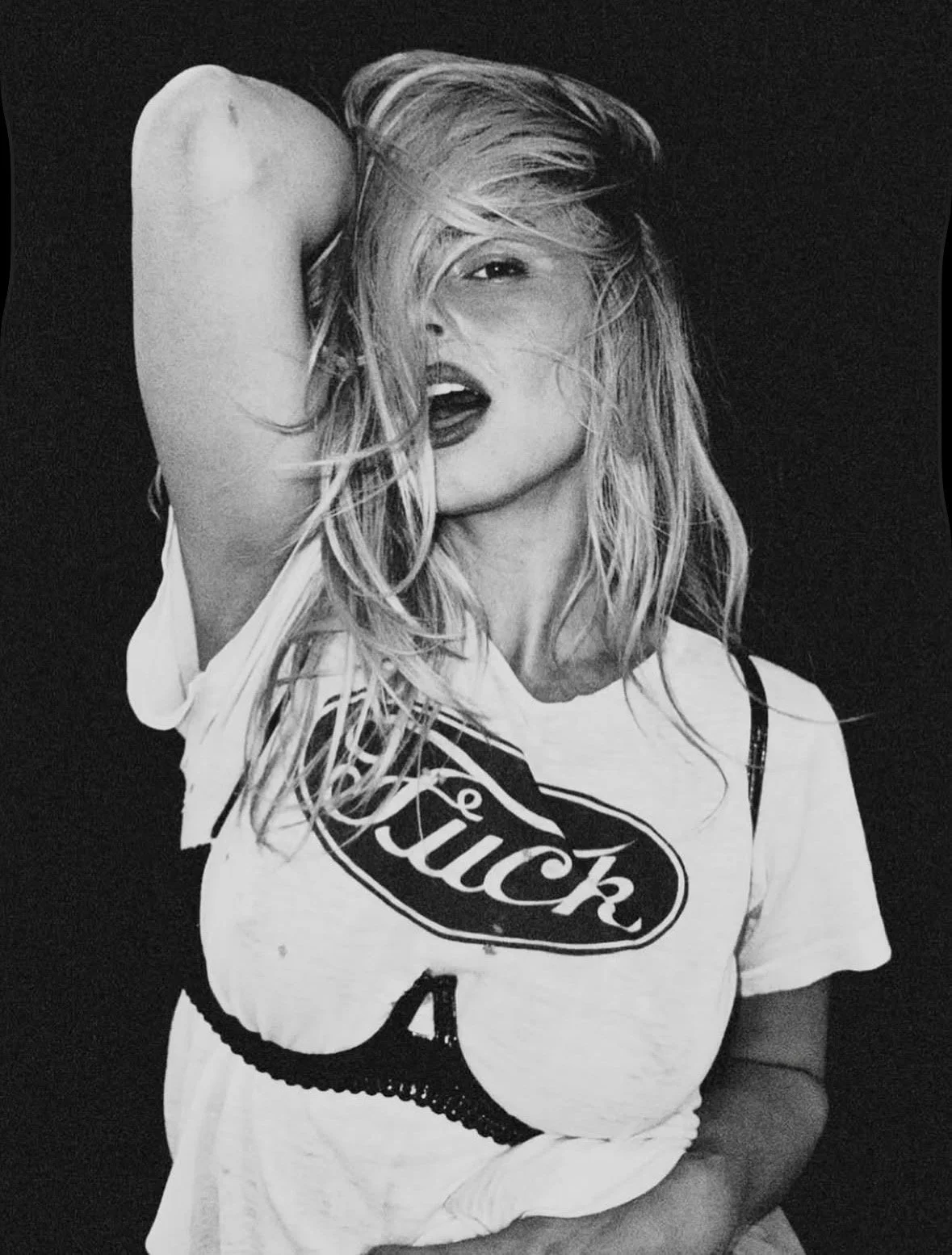Black and white photo of a woman with messy hair, wearing a t-shirt with a logo, posing with her arm over her head and her mouth slightly open.