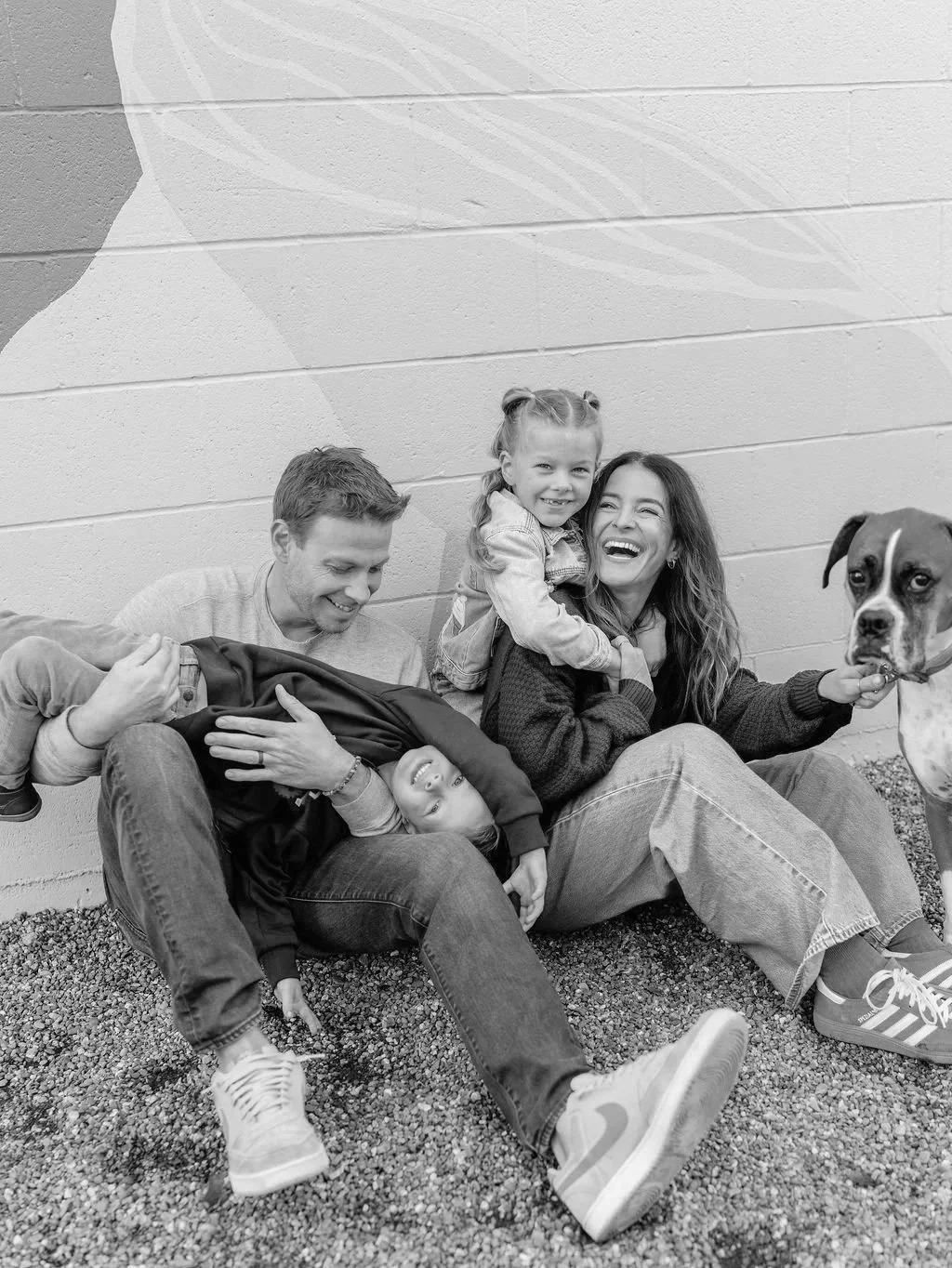 A family of five, including a man, woman, two children, and a dog, sitting and playing against a brick wall, smiling and enjoying each other's company.