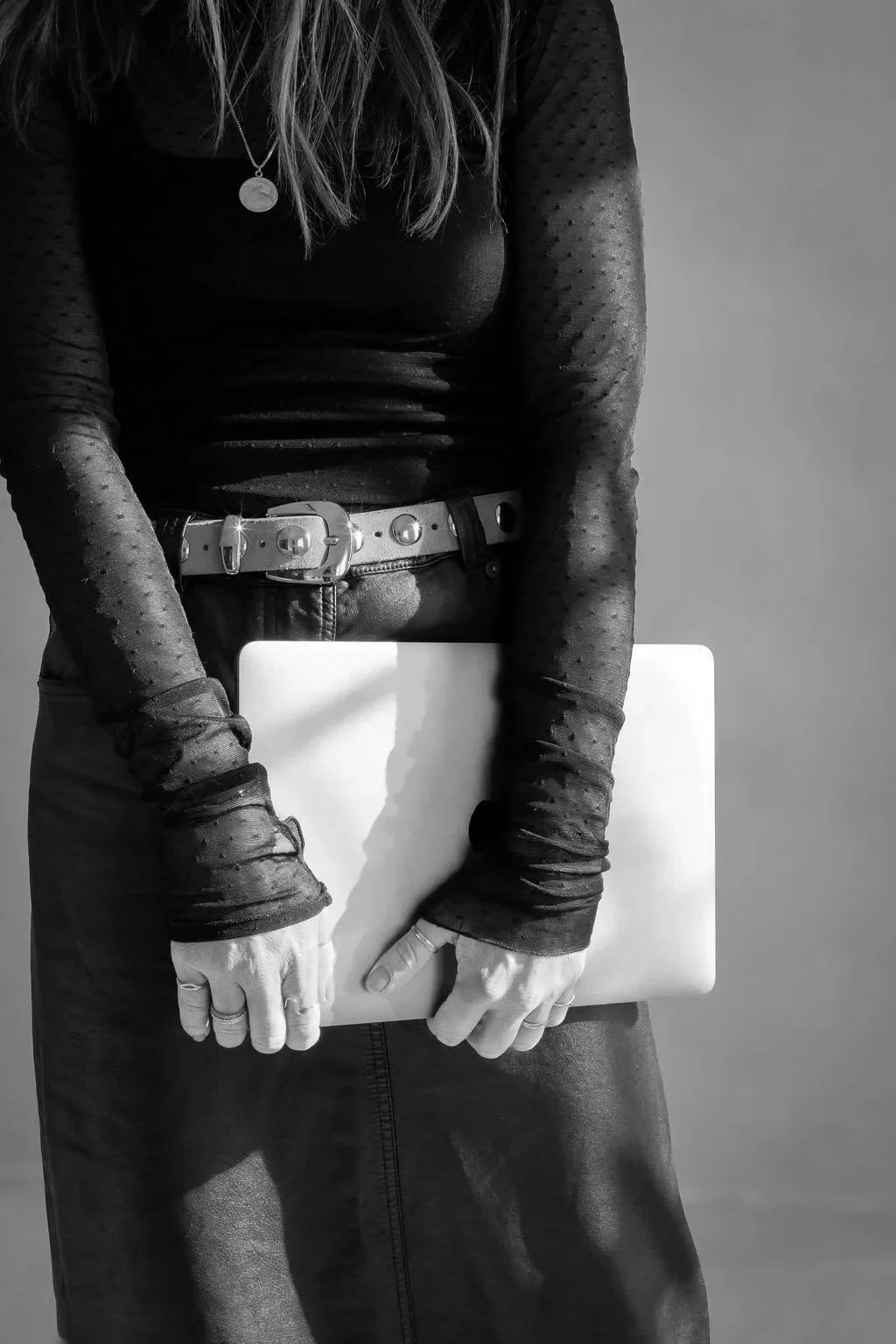 Black and white photo of a person holding a closed laptop in front of their waist, wearing a long-sleeve sheer polka-dot top and a belt with circular embellishments.
