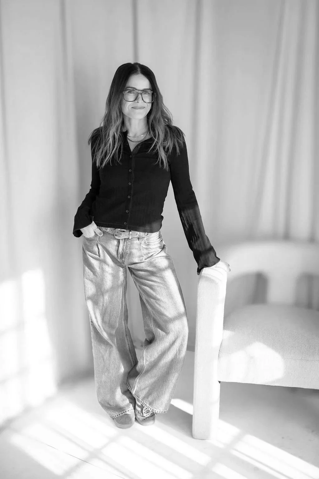 A woman with long wavy hair, glasses, and a black shirt stands indoors against a backdrop of curtains. She is smiling slightly, with her left hand in her pocket and her right hand resting on a chair.