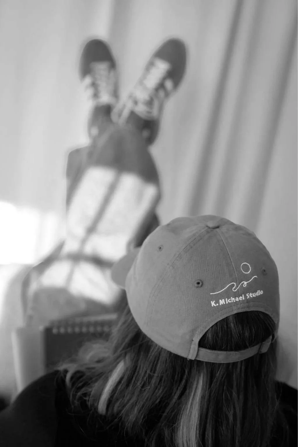 A person with long hair wearing a cap with the logo 'K. Michael Studio' is in the foreground, facing away. In the background, a projection shows someone in sneakers and shorts lying on their back on a skateboard.