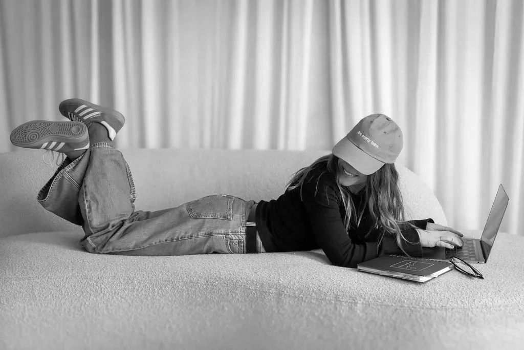 A woman lying on her stomach on a sofa, wearing a cap, working on a laptop, with a notebook and glasses nearby.