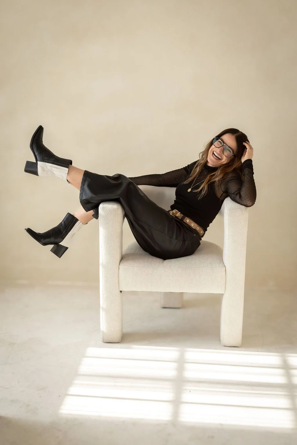 A woman sitting on a chair with her legs extended, wearing black leather pants, black boots with block heels, a black top, glasses, and smiling, against a plain beige background.
