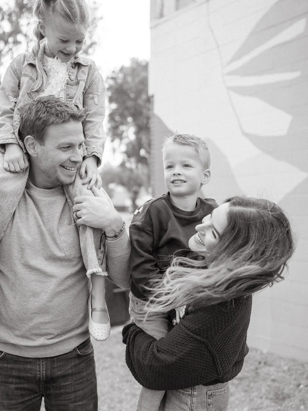 A smiling woman holding a young boy, with a man giving a piggyback ride a girl on his shoulders, outdoors near a building with graphic art.