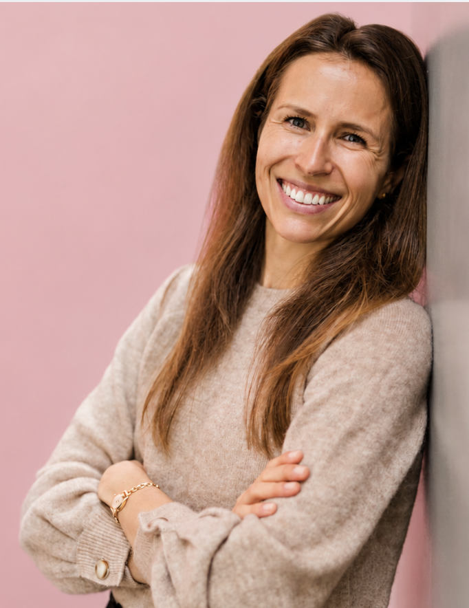A woman with long, light brown hair, wearing makeup and earrings, smiling softly at the camera in a dimly lit environment.