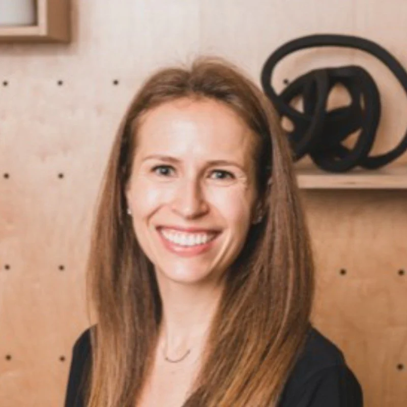 A woman with long brown hair smiling indoors with a wooden wall and a black coiled object in the background.