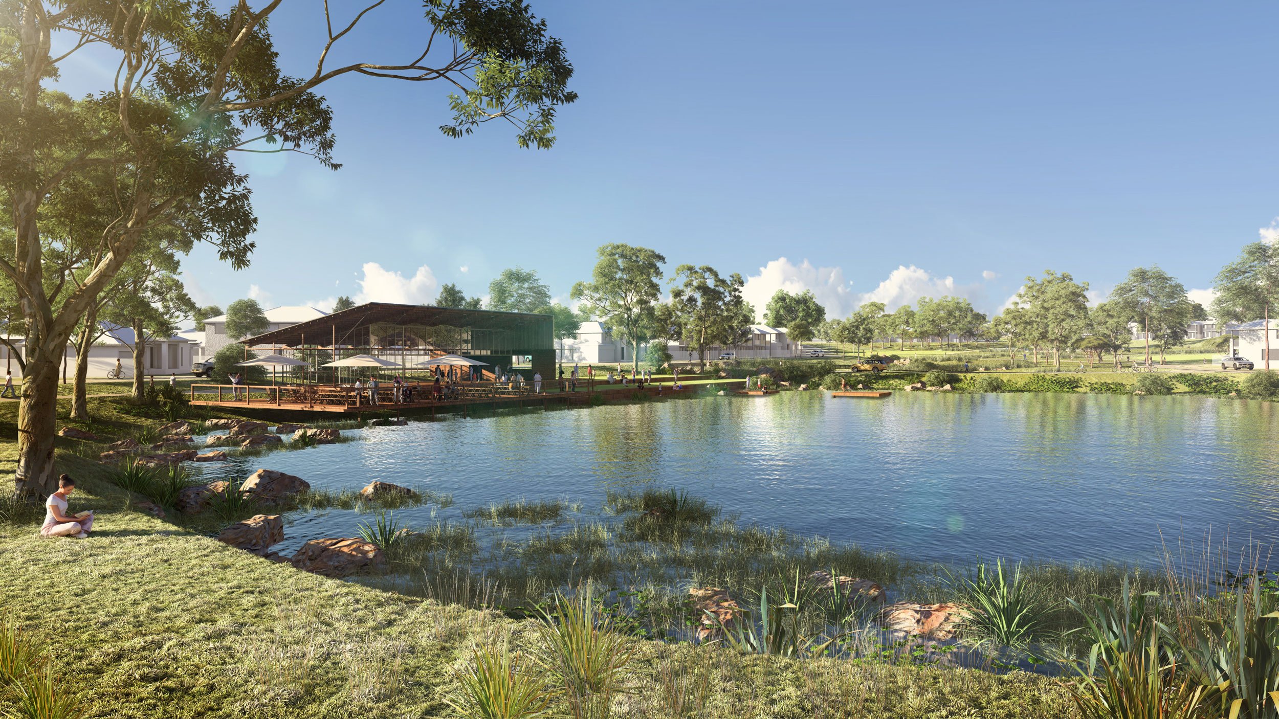 A scenic modern park features a body of water in the foreground, with people relaxing and enjoying the view. A pavilion with umbrellas and seating is set near the water, surrounded by trees and grassy areas. In the background, there are residential buildings and a road with cars and bicycles, under a clear blue sky with scattered clouds.