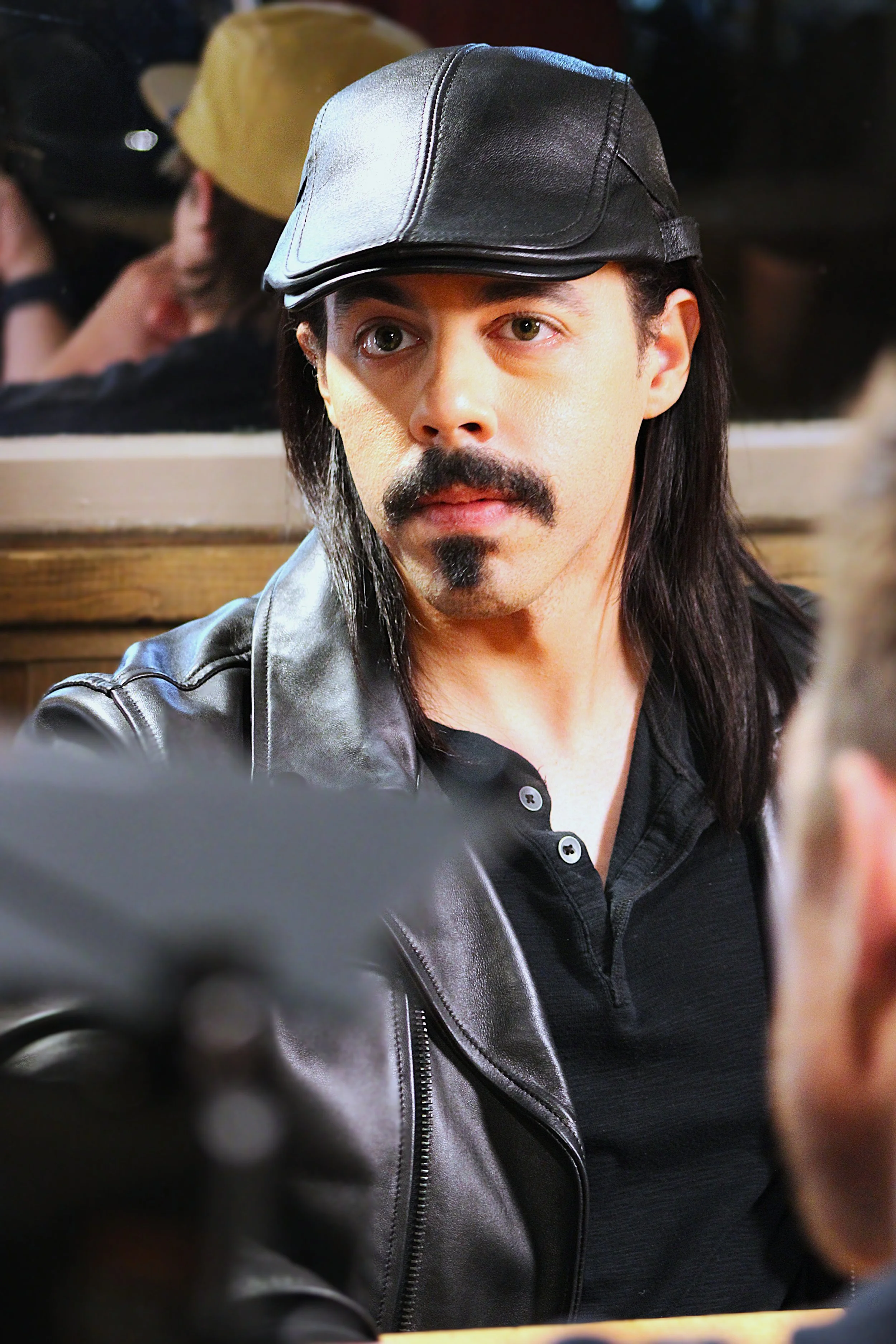 A man with long black hair, a mustache, and a goatee wearing a black leather cap and black leather jacket, sitting at a table in a busy indoor setting.