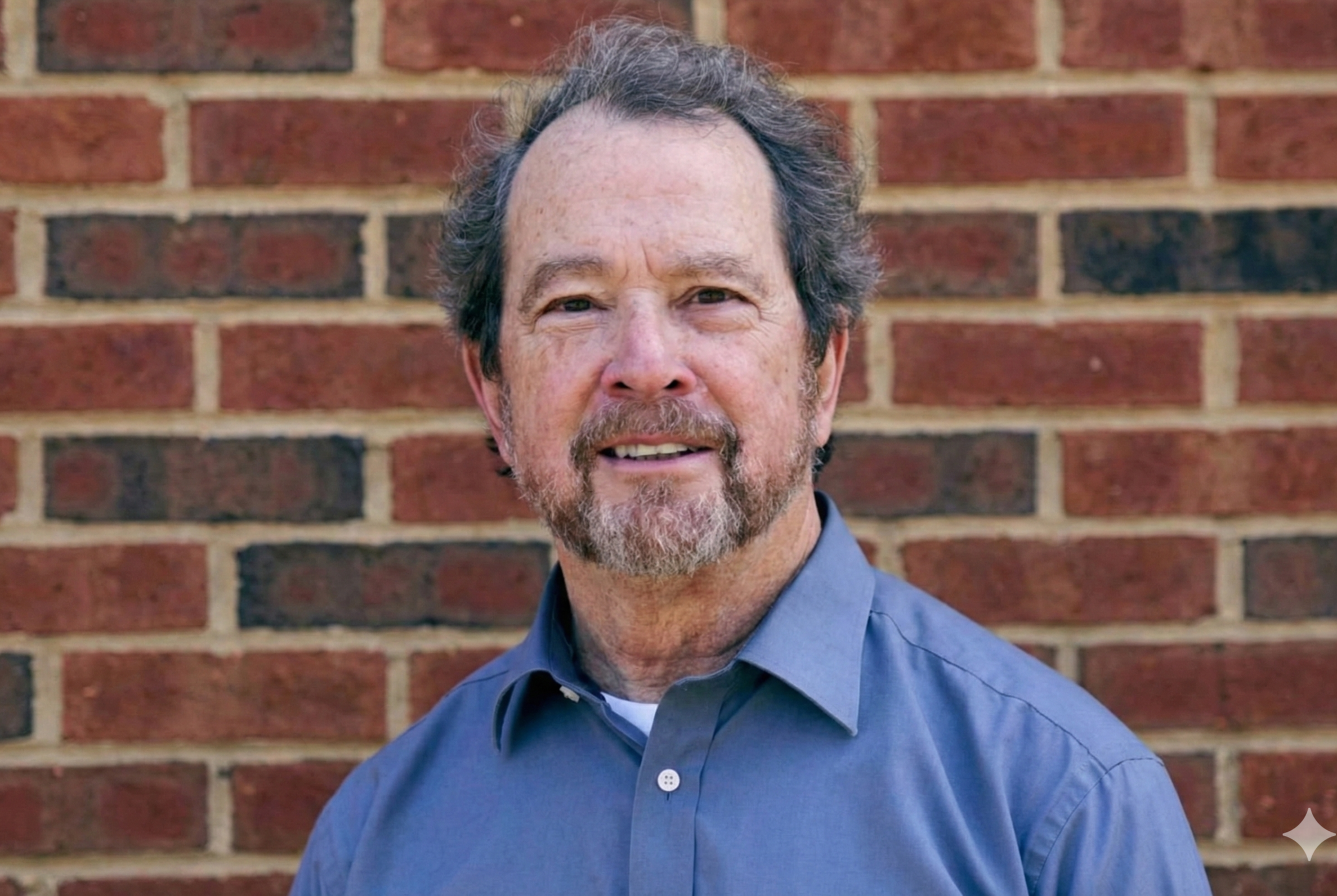 Man with beard and mustache wearing a blue shirt, standing outdoors with trees in the background.