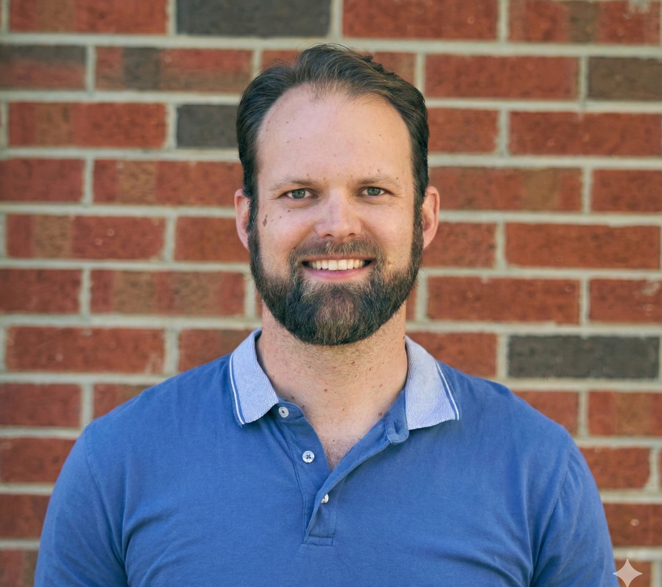 Man in a blue shirt smiling outdoors with a forest background.