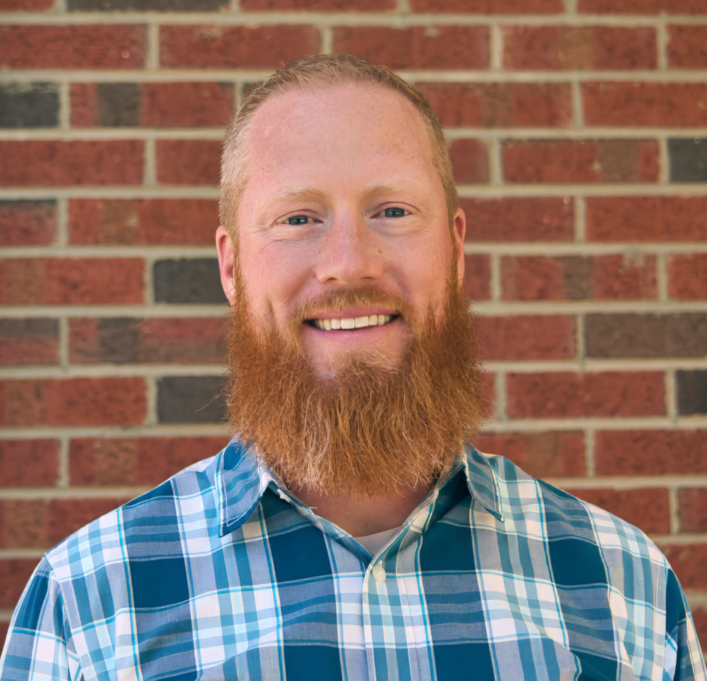 Smiling man with a beard wearing a plaid shirt standing against a brick wall.