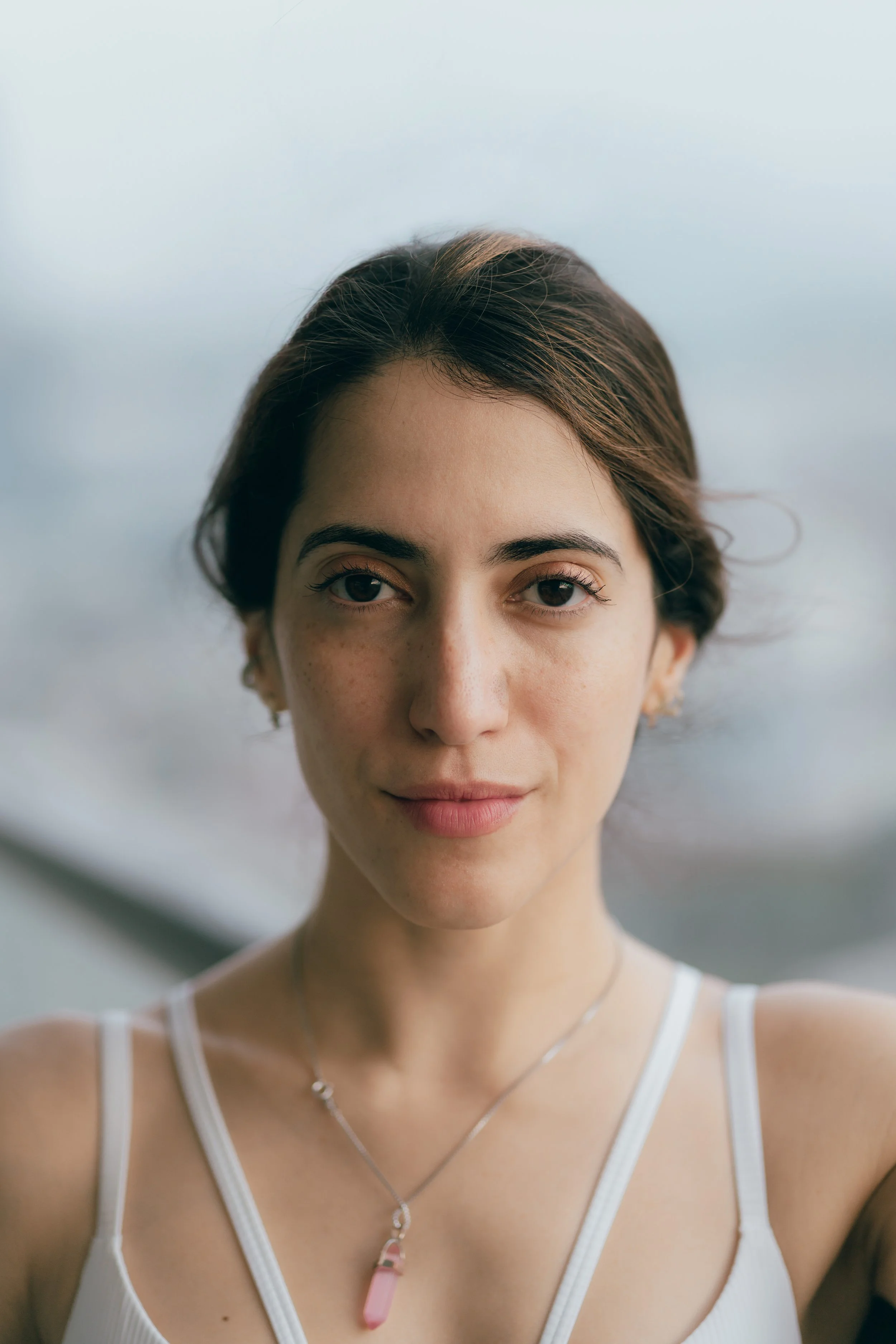 A young woman with short brown hair, light skin with freckles, and wearing a white tank top and a pink crystal pendant necklace, looking at the camera with a calm expression.