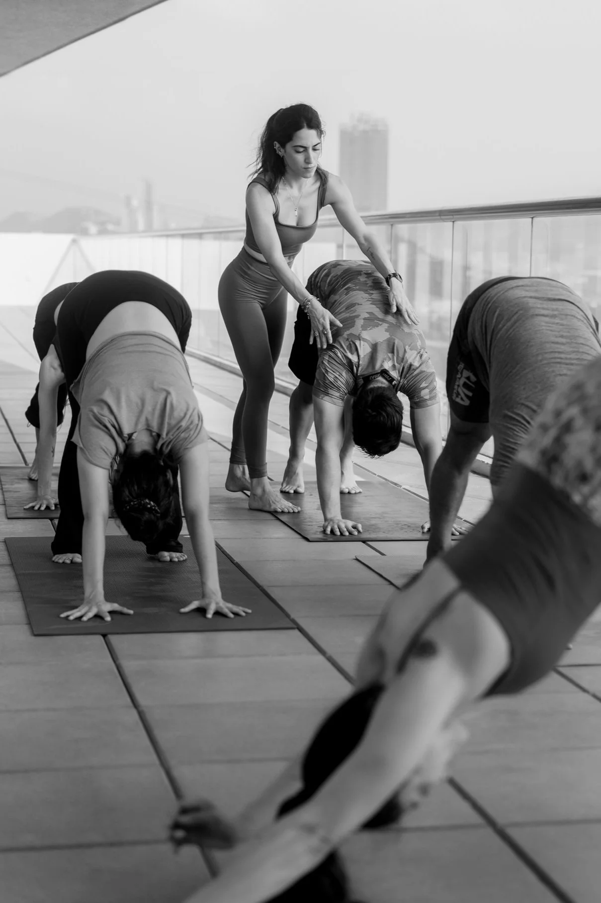 A woman instructing a yoga class on a rooftop, standing next to students in a downward dog pose, with a city skyline in the background.