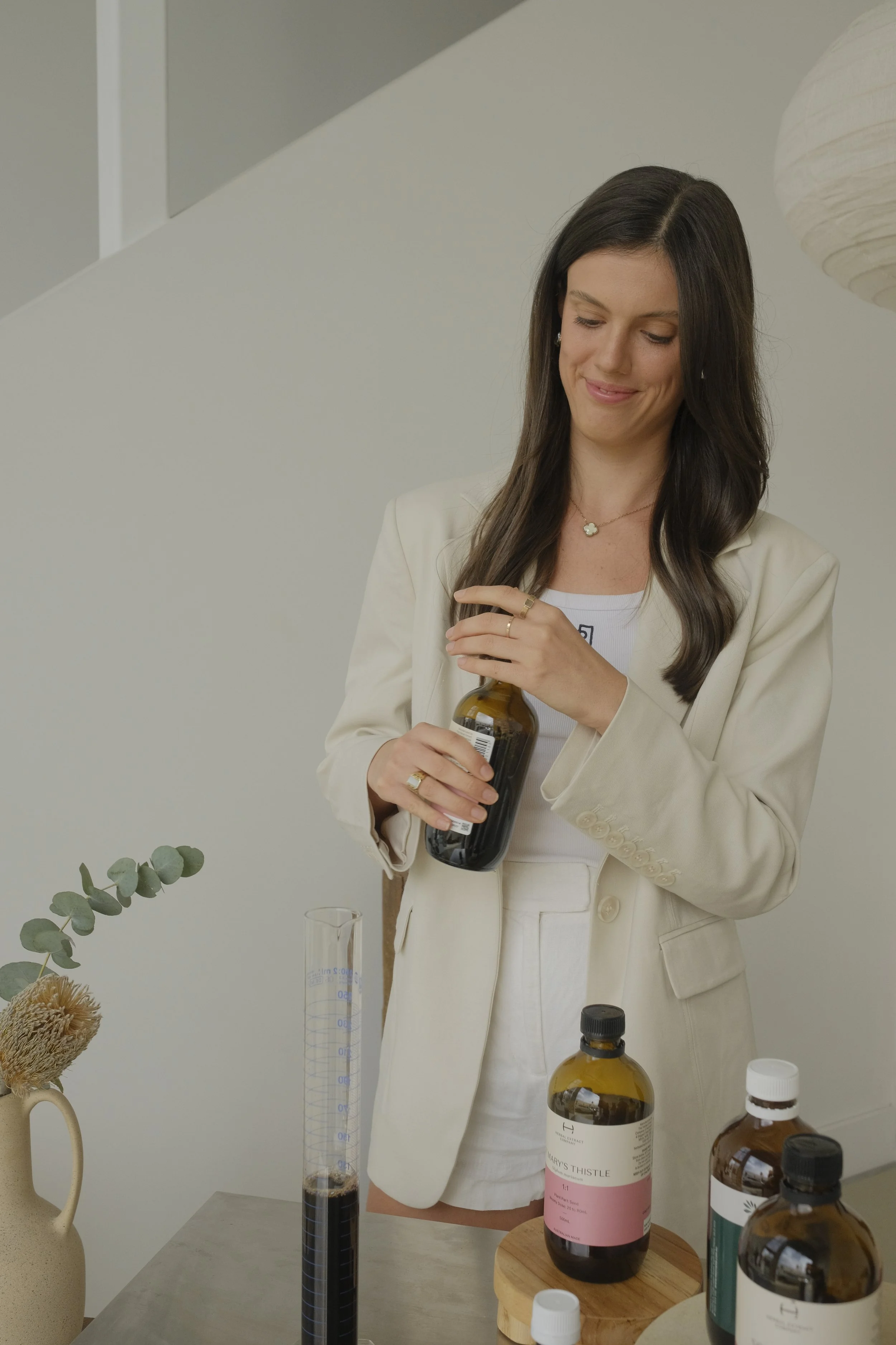 A woman in a cream-colored blazer and white outfit holds a brown bottle of herbal supplement, standing next to bottles of supplements, a beaker, and a vase with eucalyptus and dried flowers on a table.
