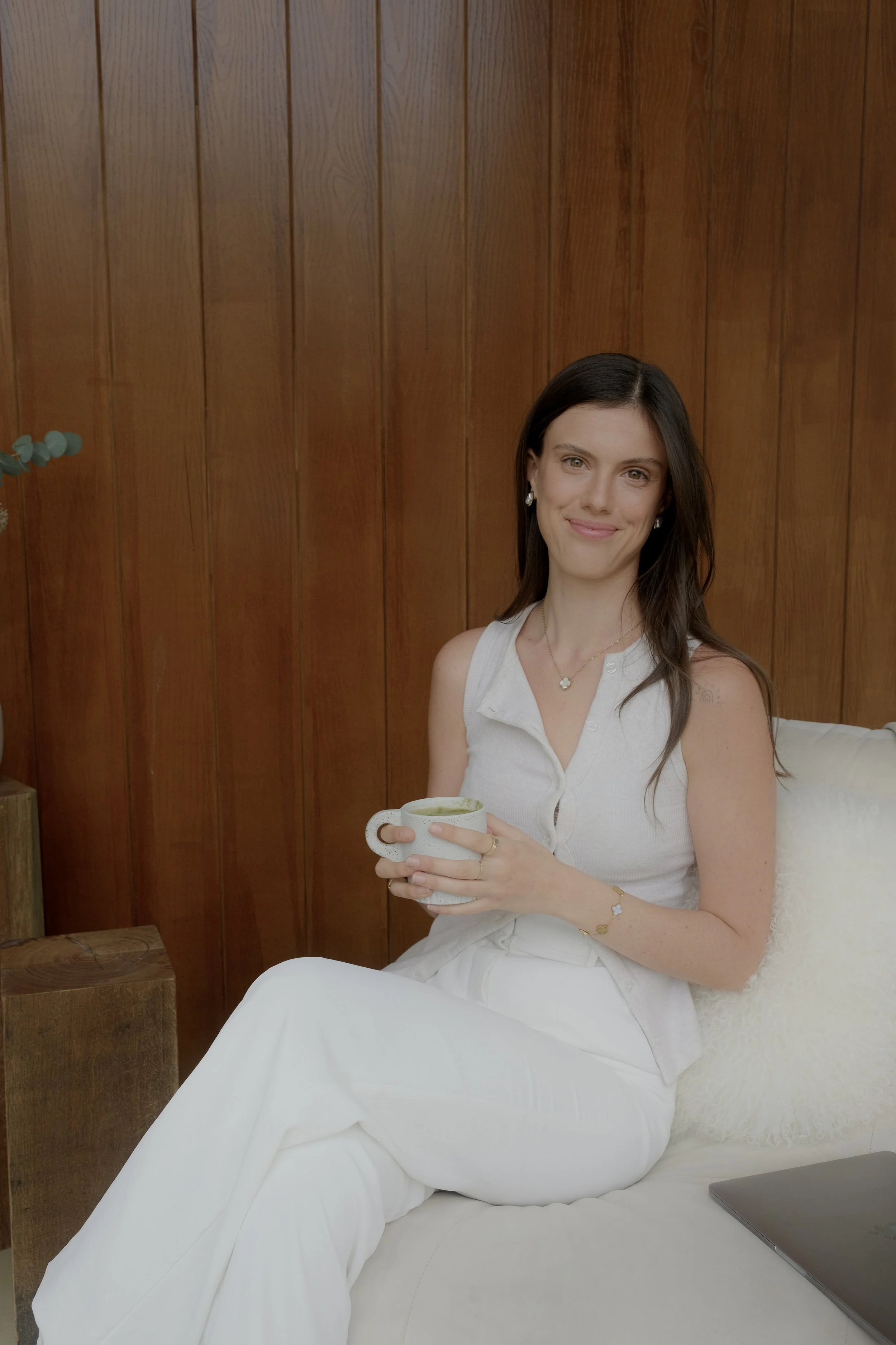 A woman with long dark hair, dressed in white, sitting on a cream-colored couch with a white pillow, holding a cup, in front of a wooden wall.