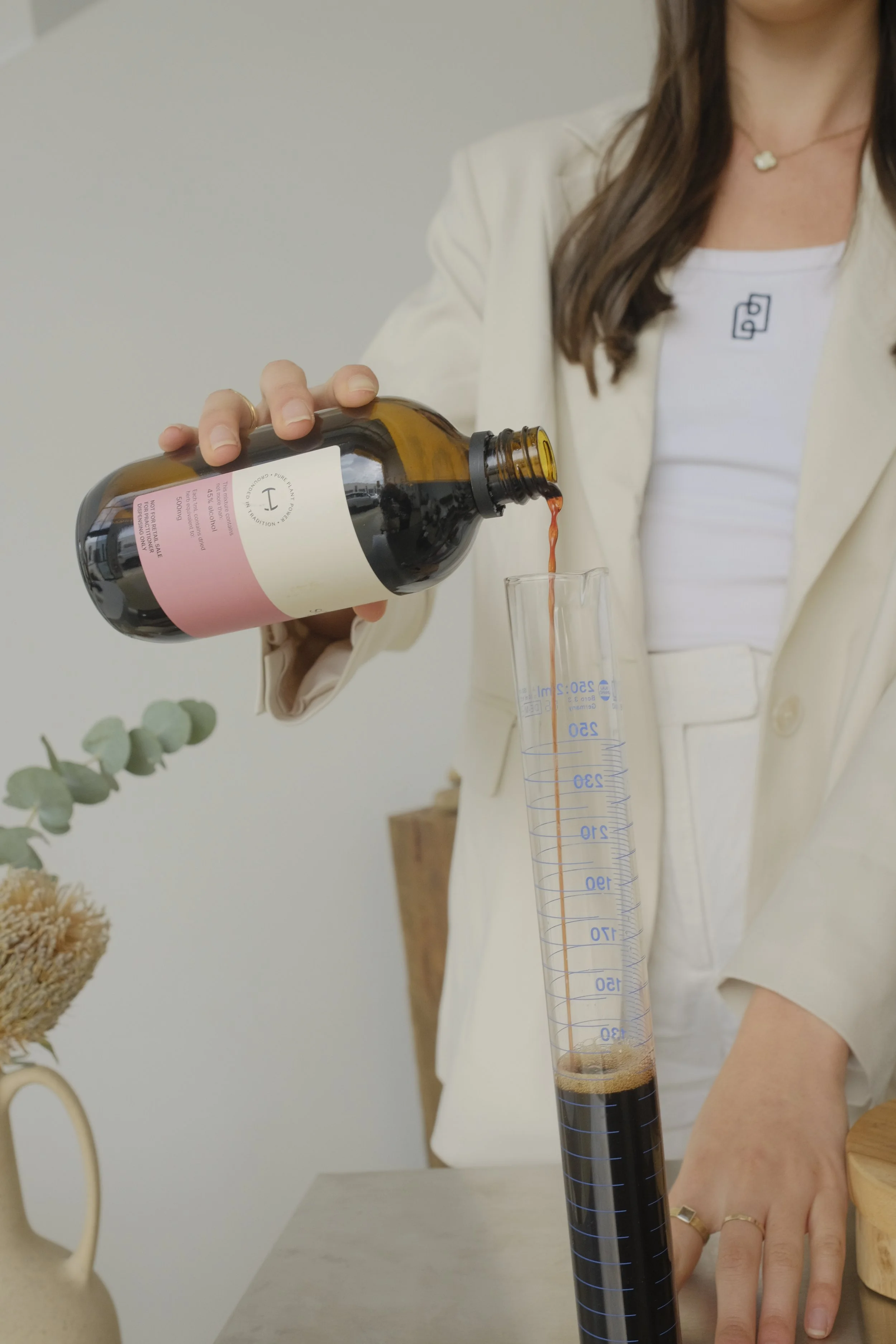 A woman in a white blazer pouring a dark liquid from a bottle into a graduated cylinder.