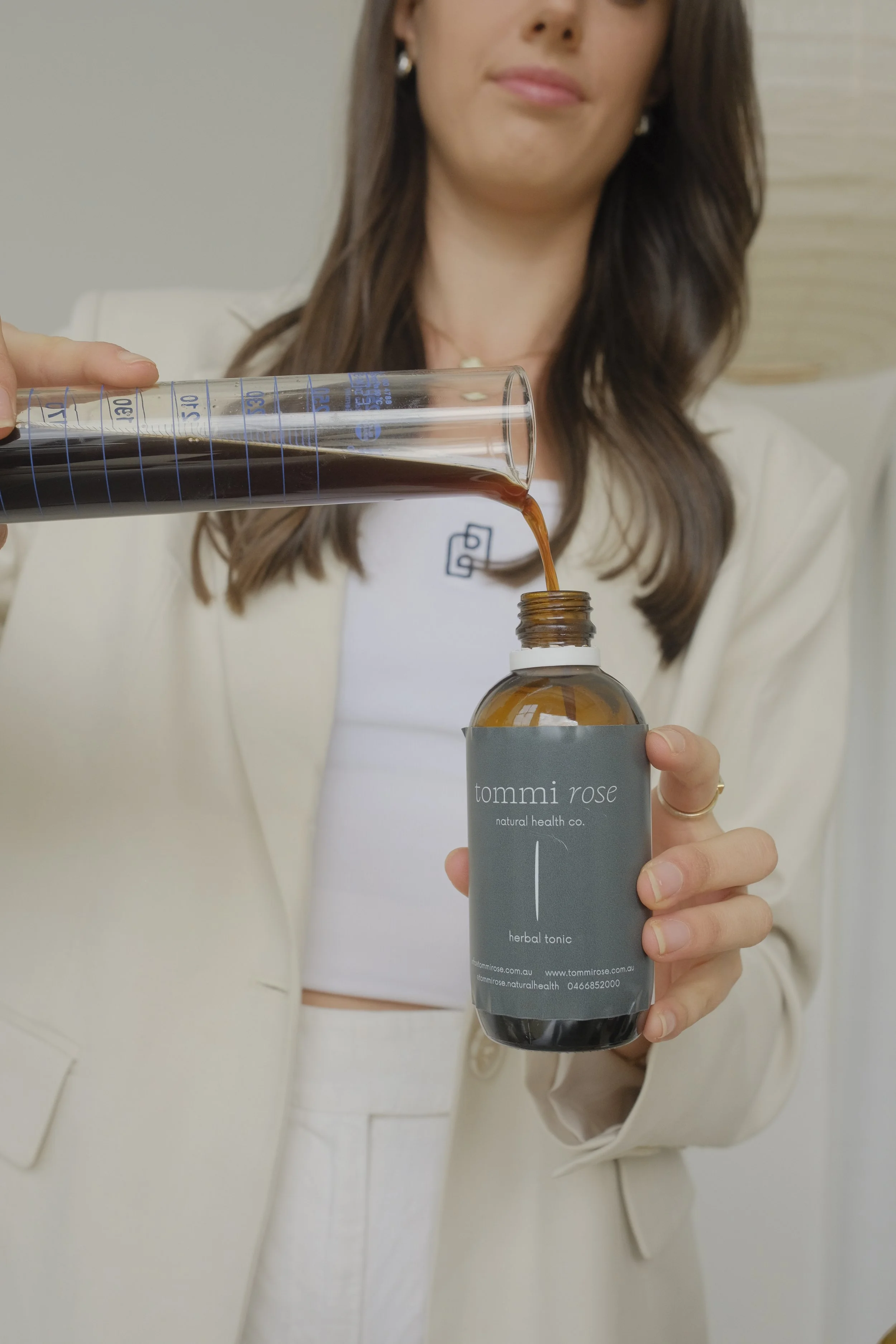 A woman wearing a cream-colored blazer and white top pouring herbal tonic from a measuring cup into a brown glass bottle with a gray label reading 'tommi rose natural health co. herbal tonic'. Naturopath near me.