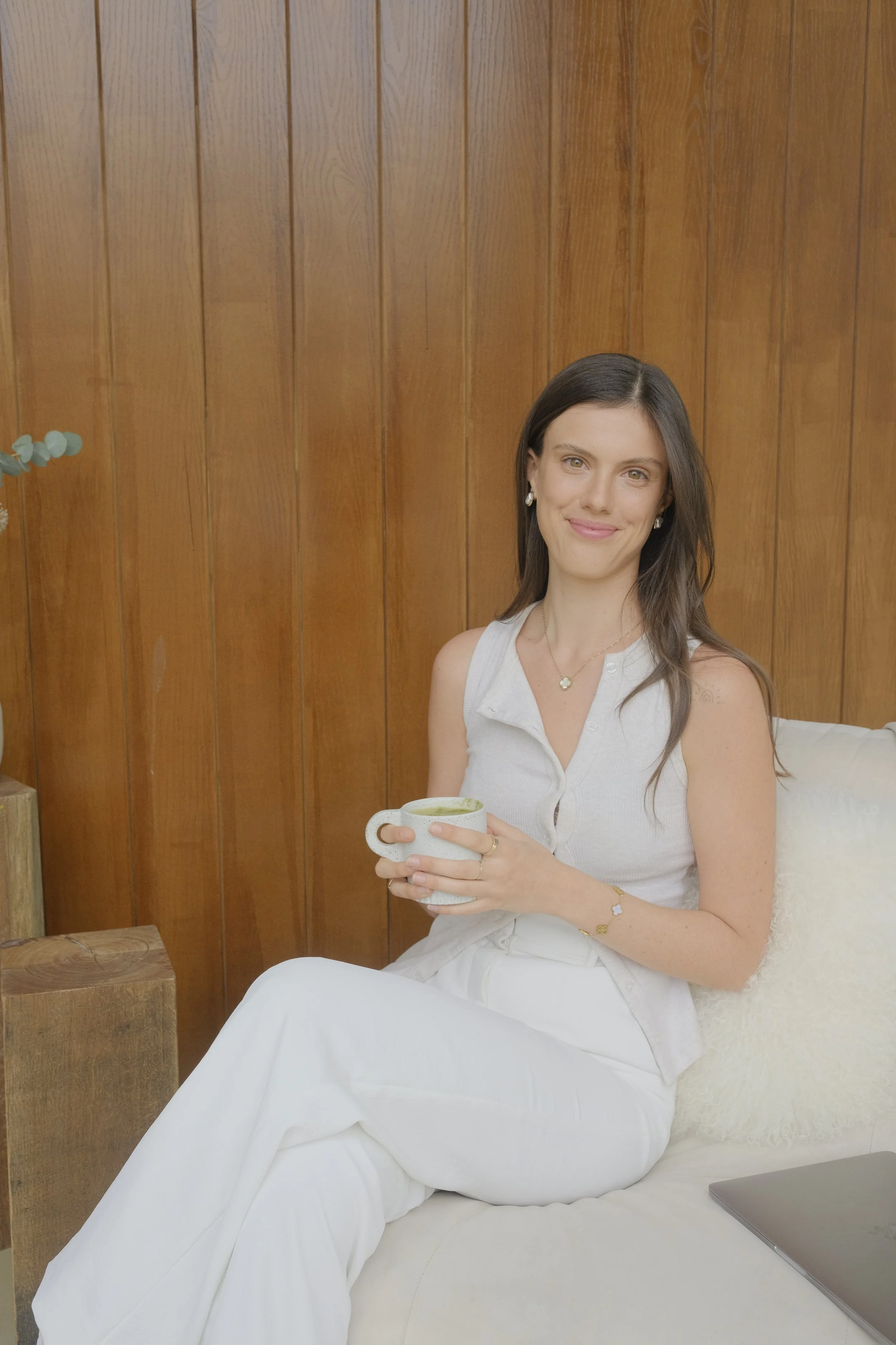 A woman with dark hair and light skin, sitting on a white sofa, holding a mug, and smiling at the camera, with a wooden wall background.