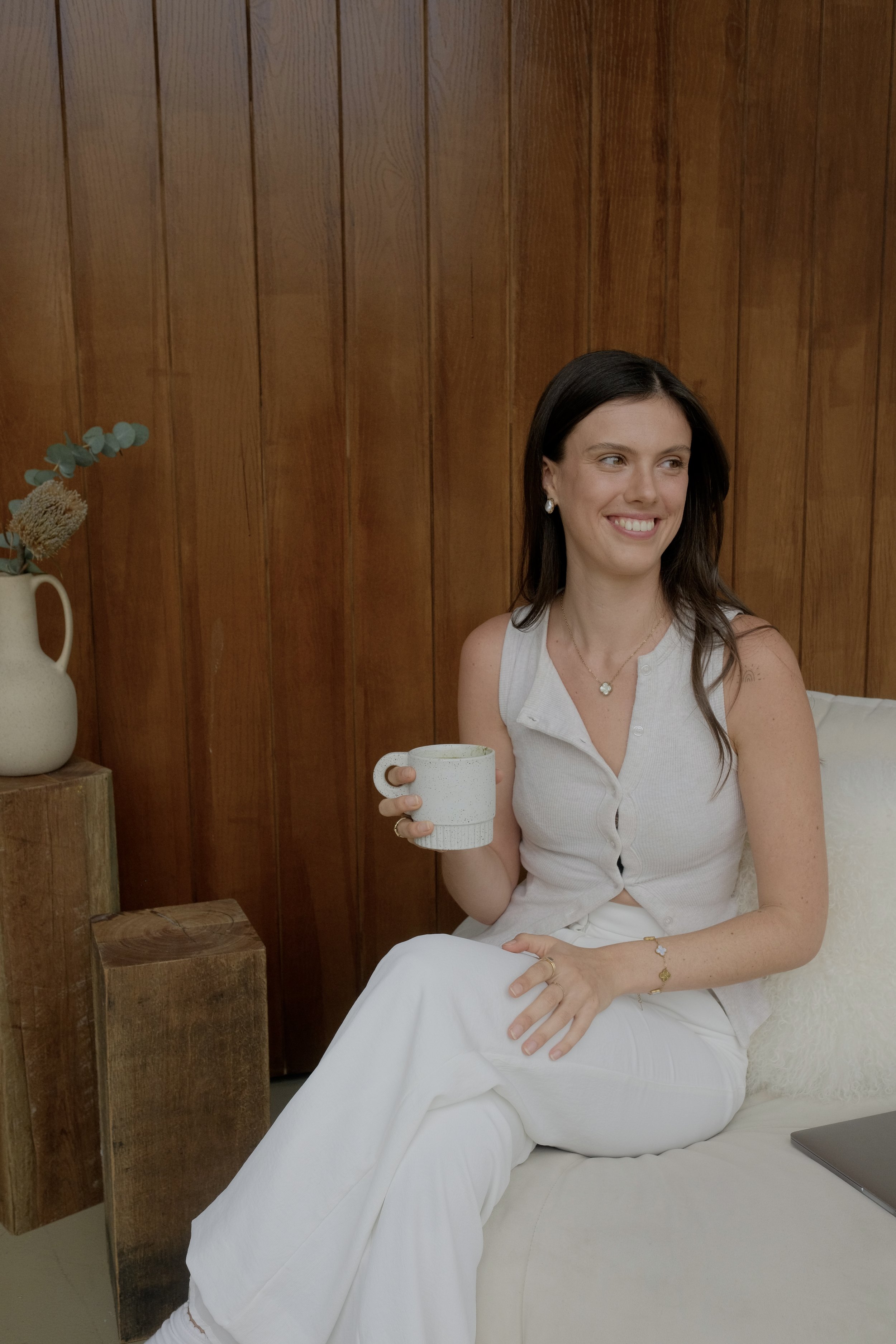 A young woman with dark hair and light skin, smiling, sitting on a cream-colored sofa, holding a mug, against a wooden wall. She wears a sleeveless button-up top, white pants, jewelry including earrings, a necklace, a bracelet, and a ring. There is a pillow behind her and a laptop on her lap. To her left, there is a wooden table with a cream vase and some greenery.