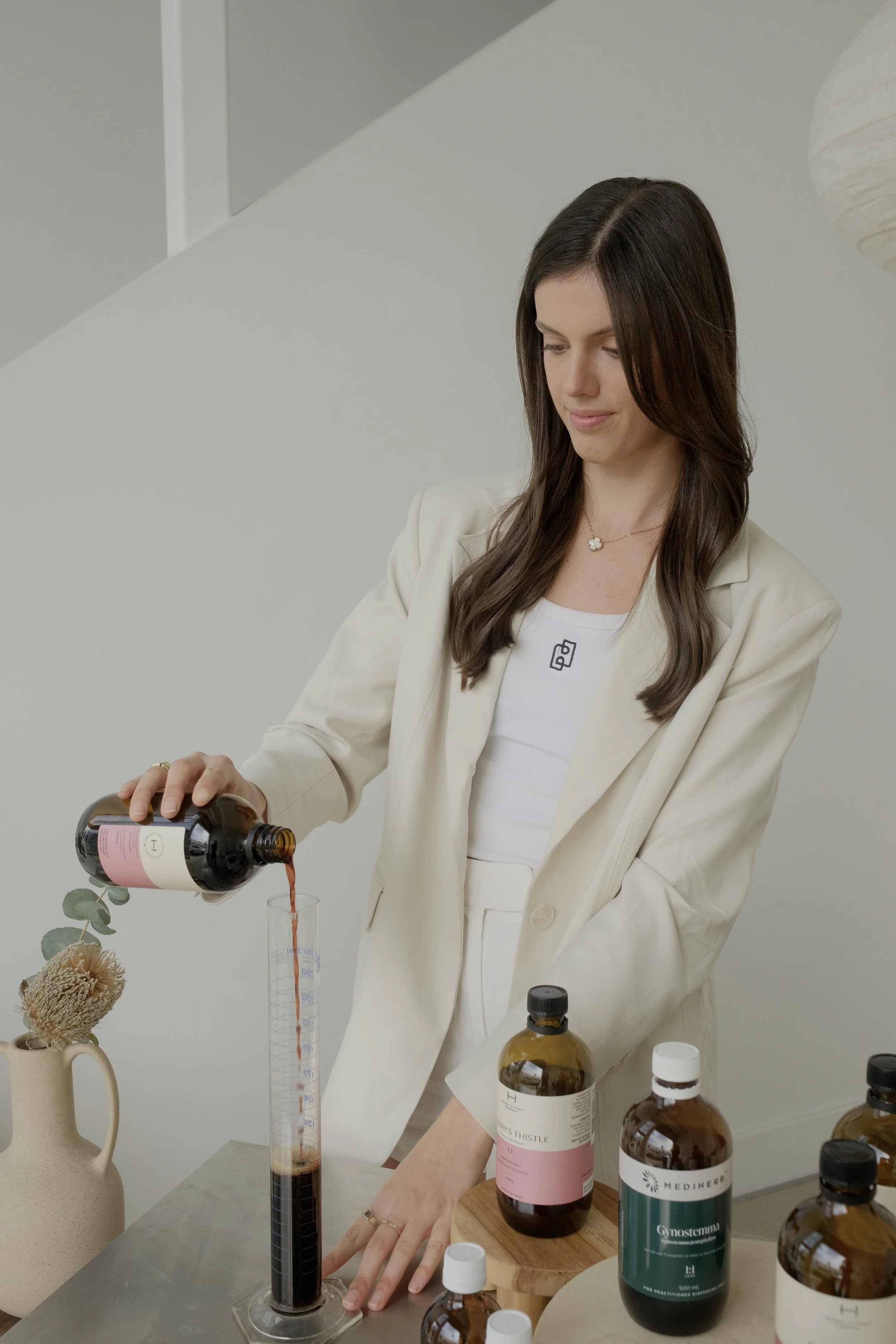 A woman in a white blazer pours a dark liquid from a brown bottle into a tall measuring cylinder. Several other brown bottles are on the table, along with a beige vase with dried flowers.