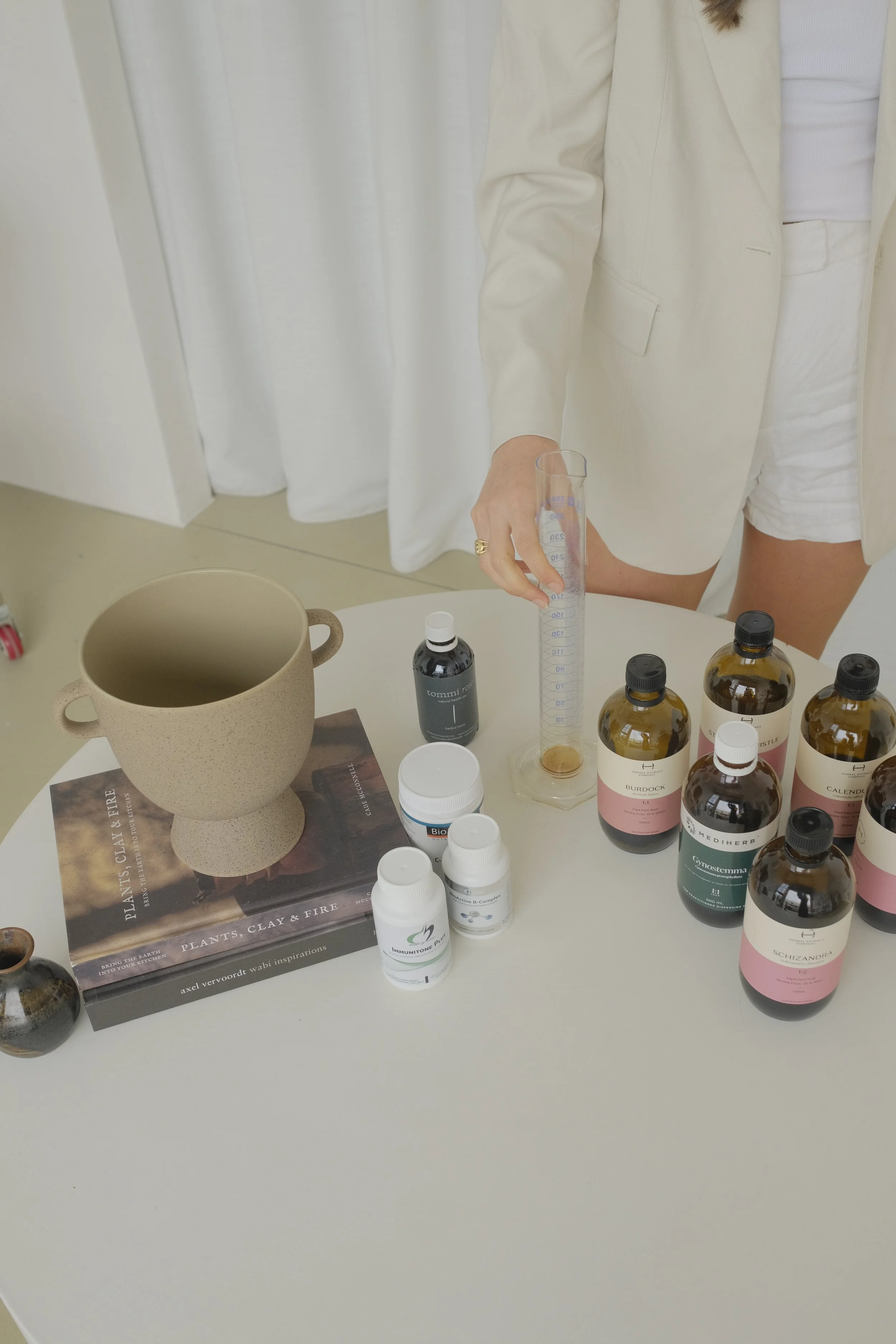 Person pouring liquid into a graduated cylinder surrounded by bottles of herbal supplements, a ceramic mug, and books on plants and fire on a white table.