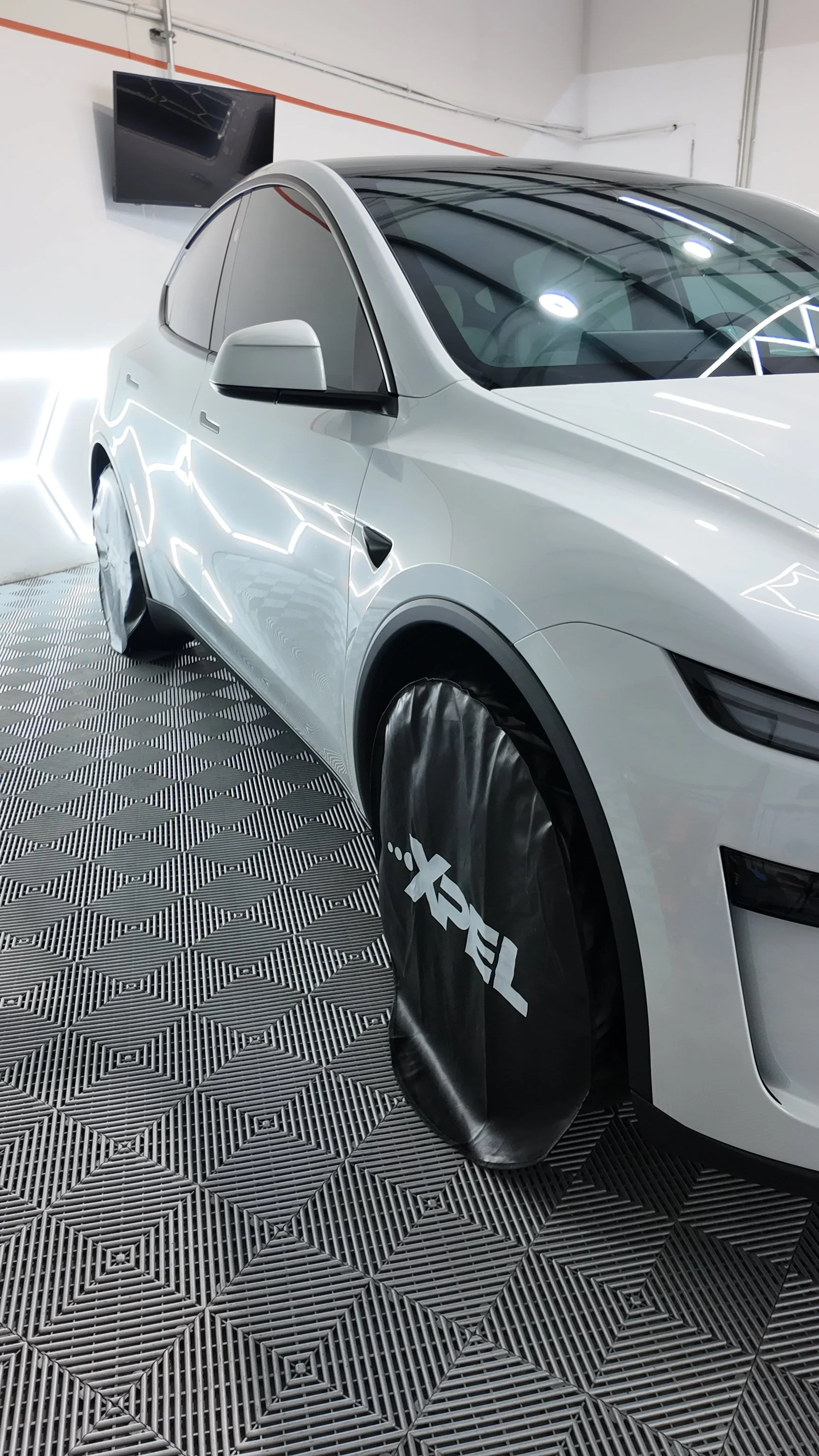 A Tesla electric vehicle is inside NextGen Auto Spa, with one wheel covered by a black protective cover featuring the Xpel logo. 