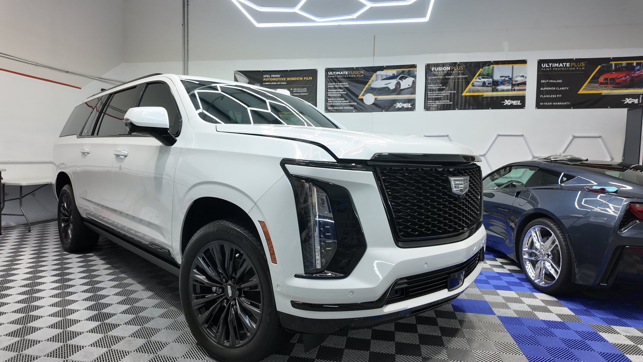 A white Cadillac SUV parked indoors with a black grille, black wheels, and modern headlights. Next to it is a gray sports car with a vinyl wrap, chrome wheels, and a low-profile design. The background features promotional banners on the wall and a patterned garage floor.