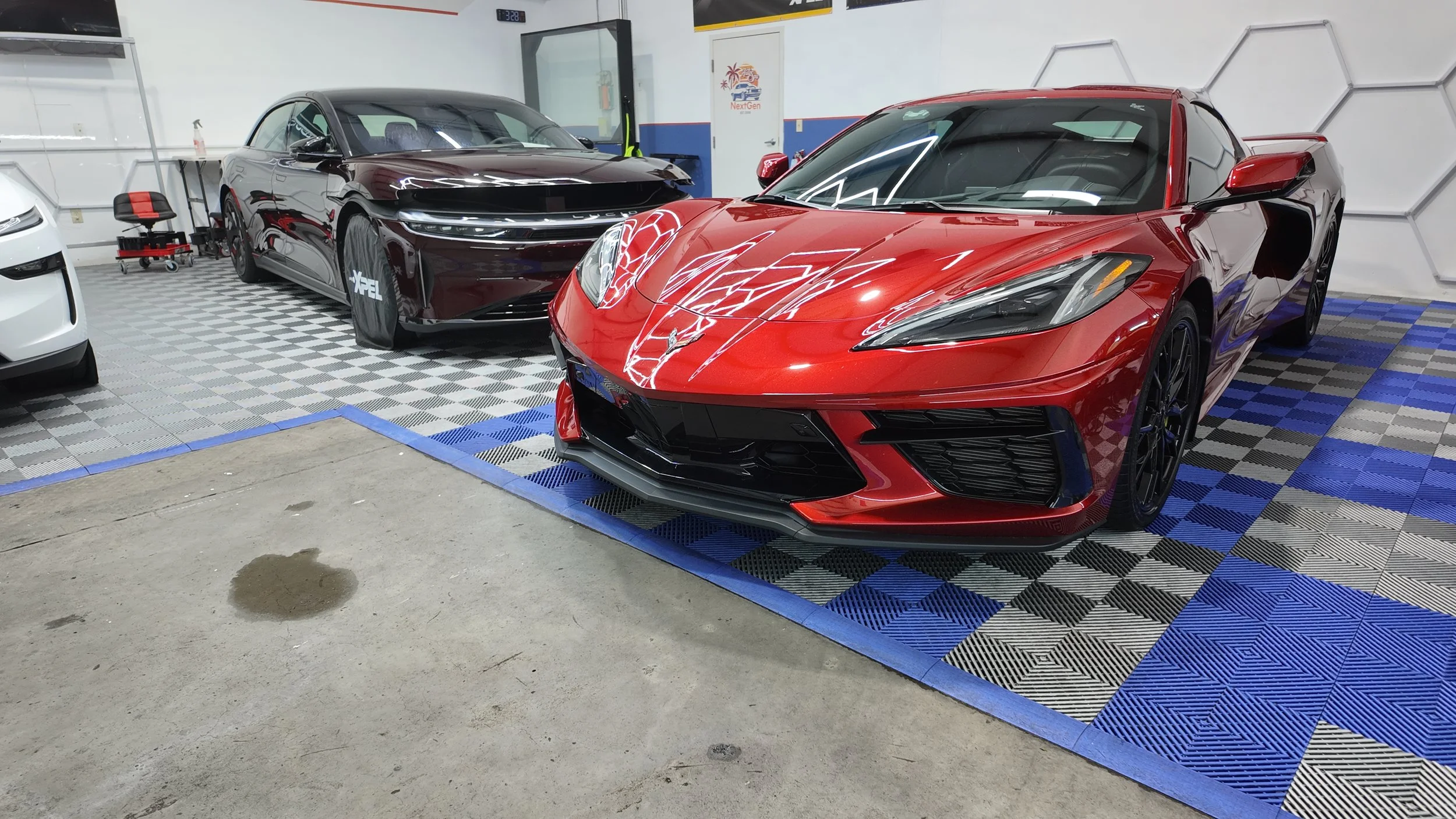 Red sports car parked inside a garage next to a black car, with the garage floor covered in checkered mats.
