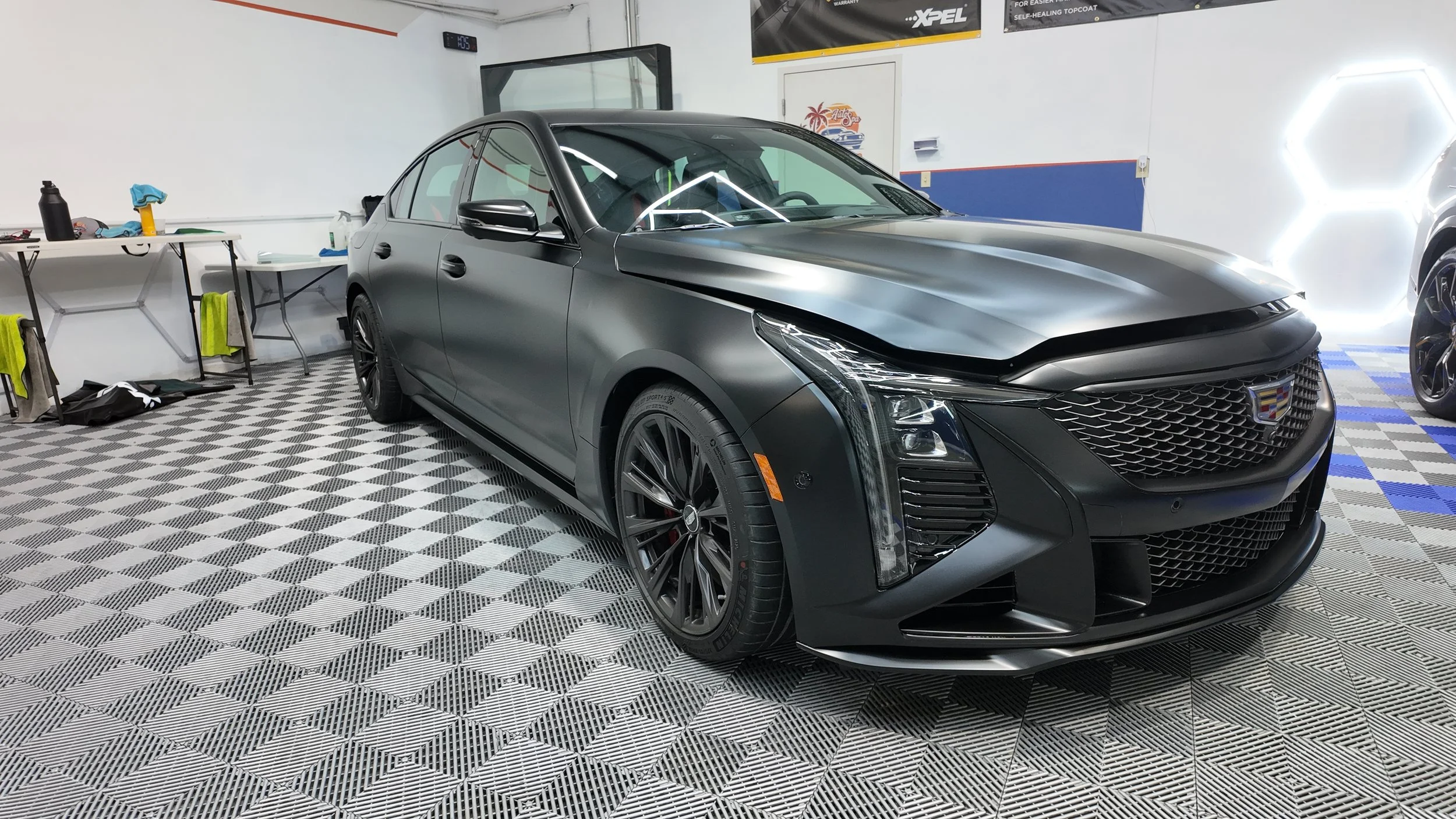 A black Cadillac sedans on a checkered garage floor with shiny black rims. The car's front hood is slightly open, and it is in a garage with work tools and colorful wall decorations.