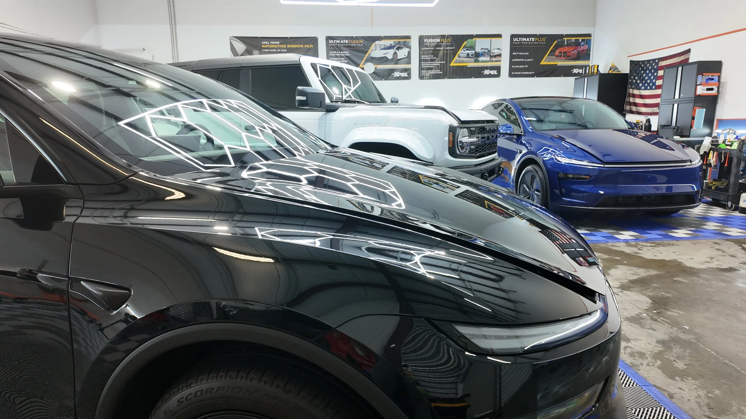 Various cars inside an auto detailing shop, including a black Tesla Model Y in the foreground, a white SUV, and a blue sedan, with posters and tools on the walls.