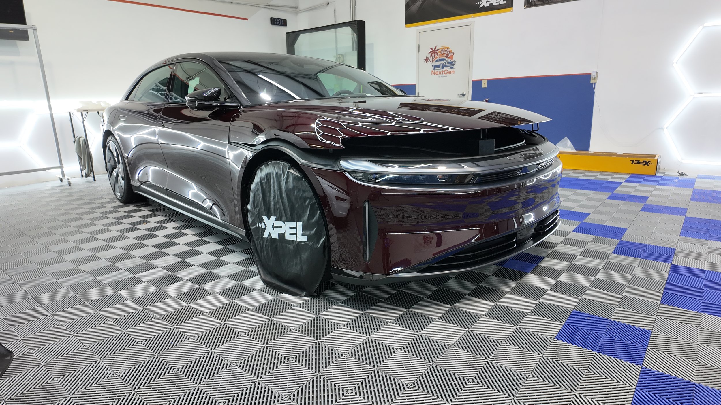 A maroon-colored Lucid electric car at NextGen Auto Spa with a cover over its front left wheel featuring the 'XPEL' logo. After paint protection film installation. 