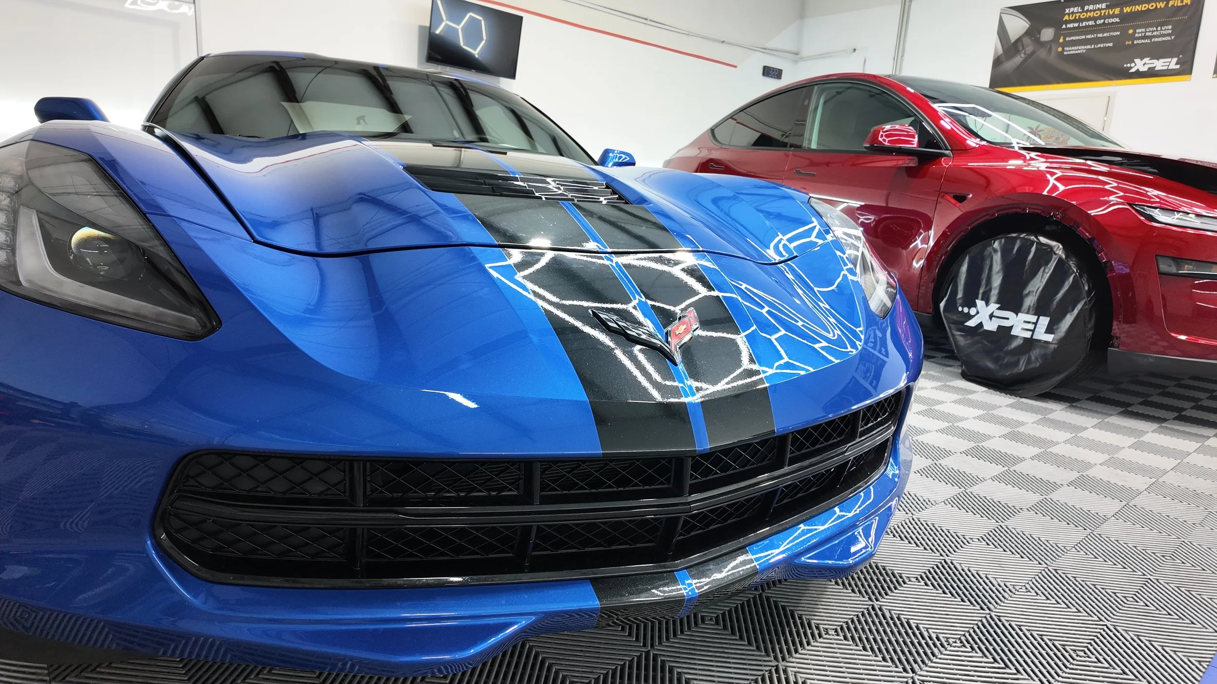 Blue Chevrolet Corvette sports car with black racing stripes next to a red Tesla Model S in a showroom.