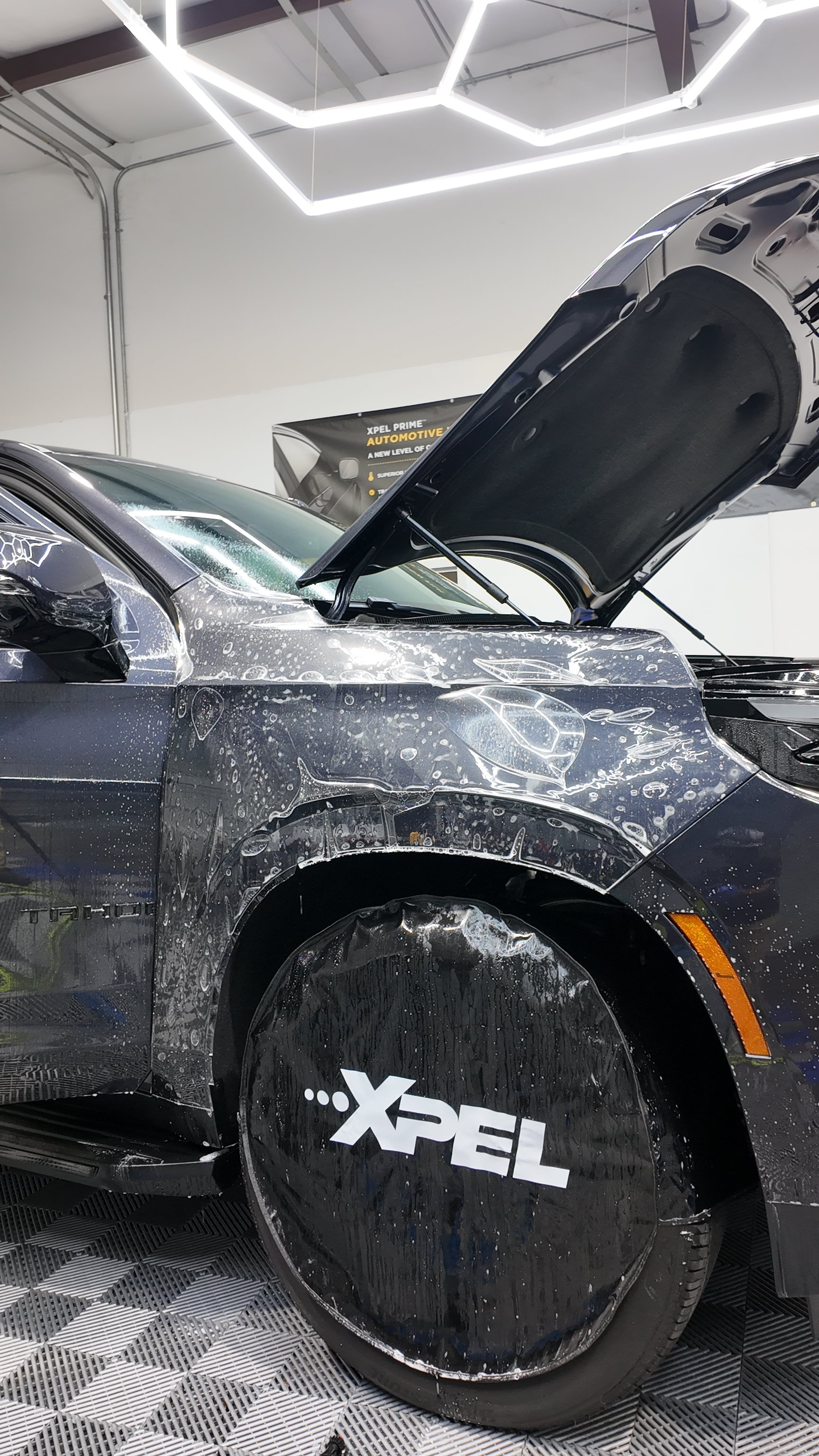 A black car with a partially open hood, covered in soap suds and water, in a garage or detailing center. The car's wheel is covered with a black cover labeled 'XPEL'. Overhead lighting is visible.