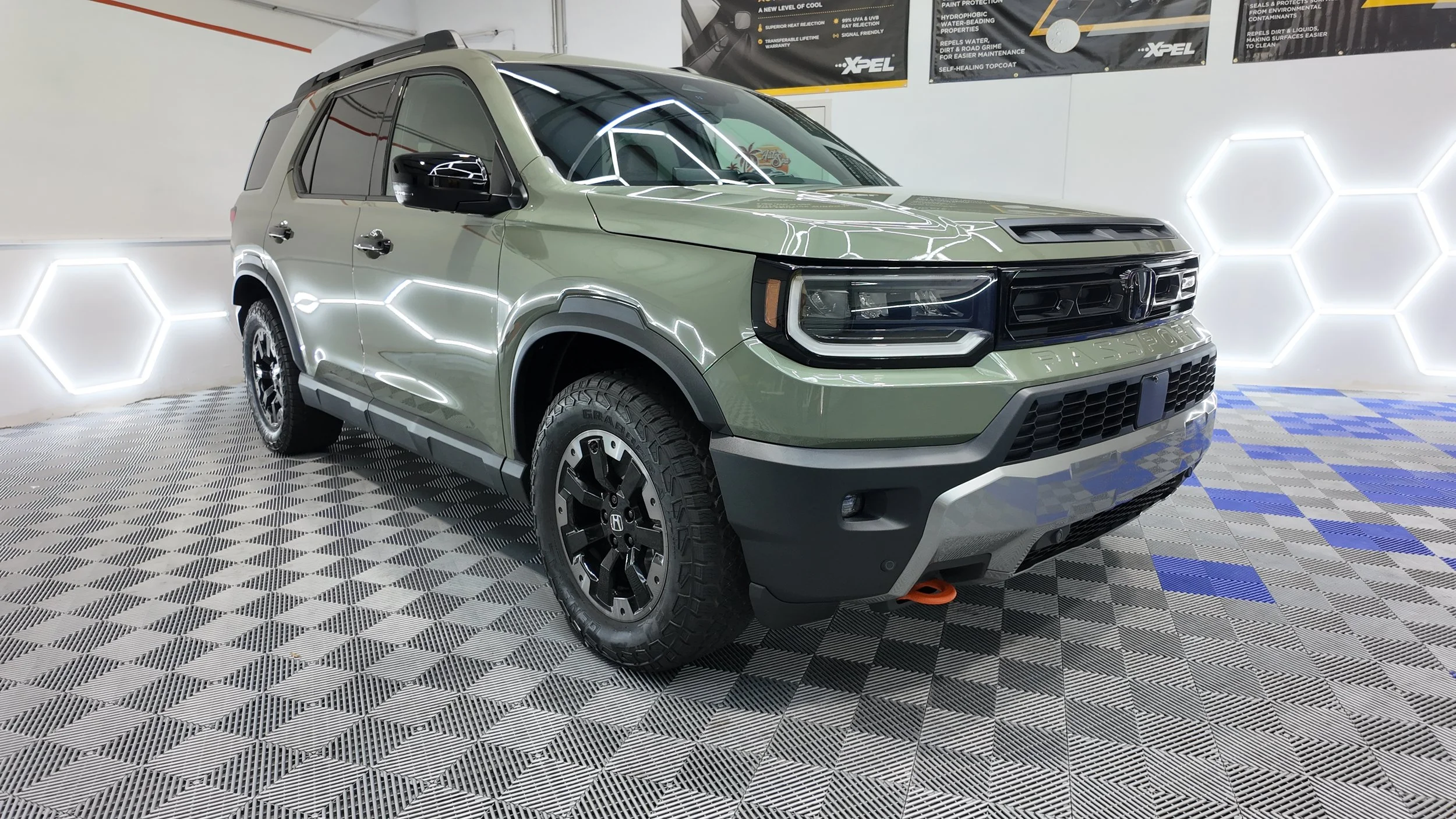 A black car with a partially open hood, covered in soap suds and water, in a garage or detailing center. The car's wheel is covered with a black cover labeled 'XPEL'. Overhead lighting is visible.