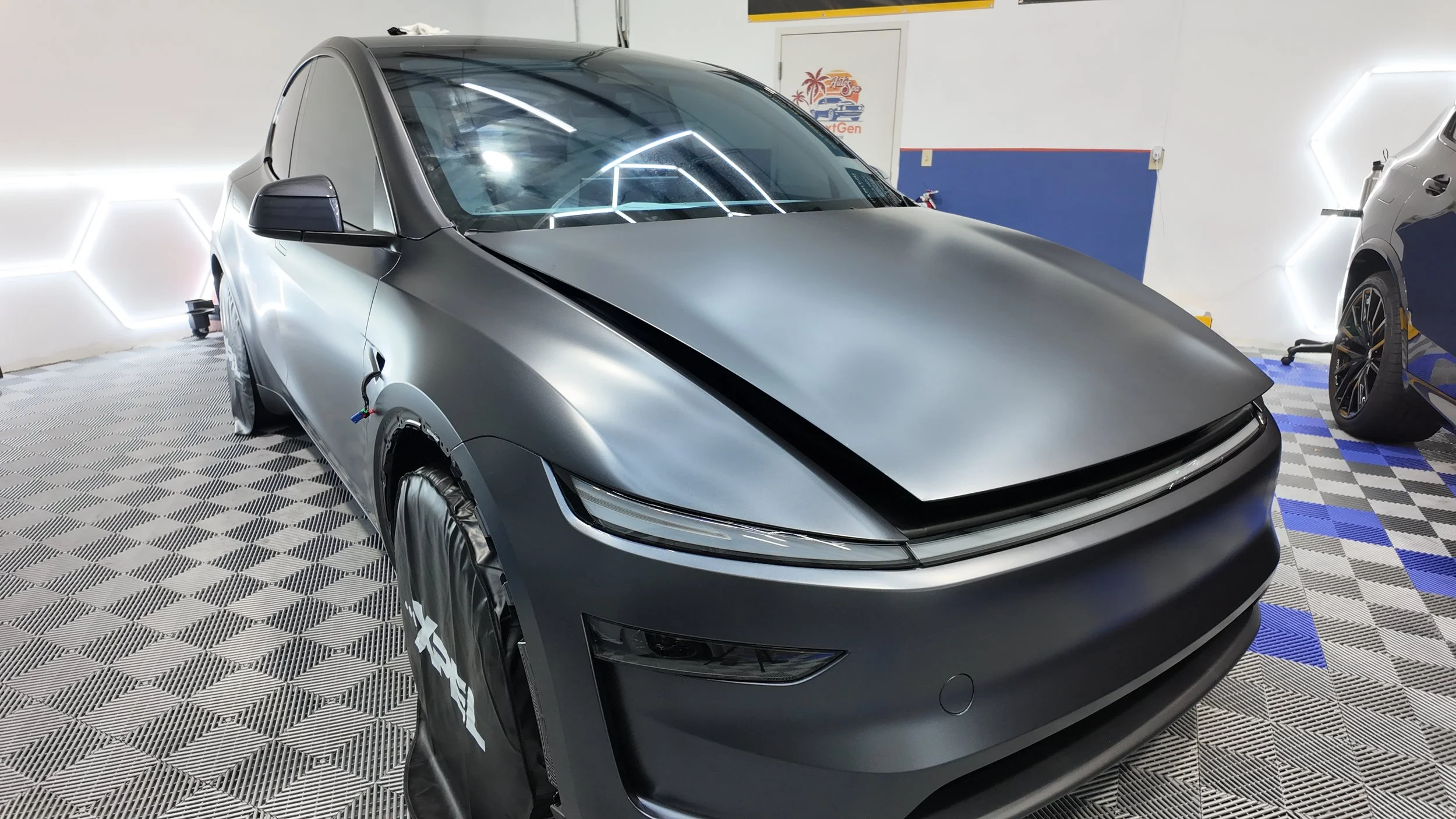 A matte gray electric car undergoing repairs in a workshop, with front wheels covered and the hood slightly open.