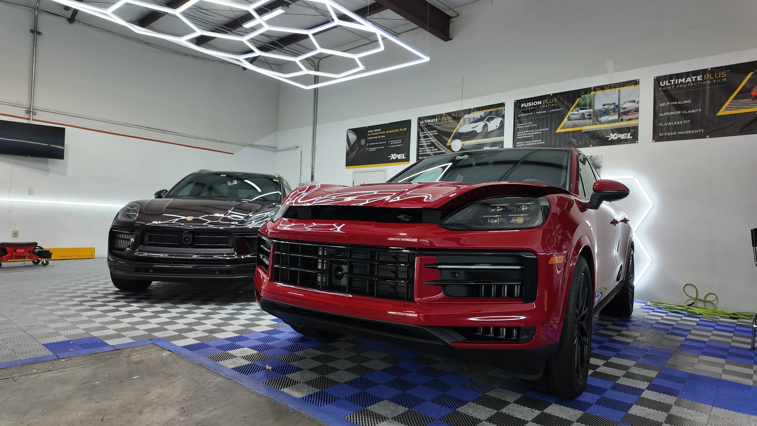 Two luxury cars, one red and one black, inside a well-lit automotive detailing garage with illuminated geometric ceiling lights and posters on the wall.